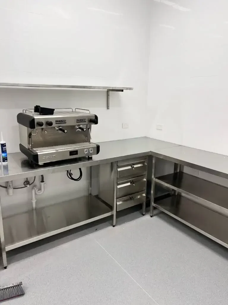 Empty commercial kitchen with stainless steel countertops, shelves, and a dual espresso machine on the left side.