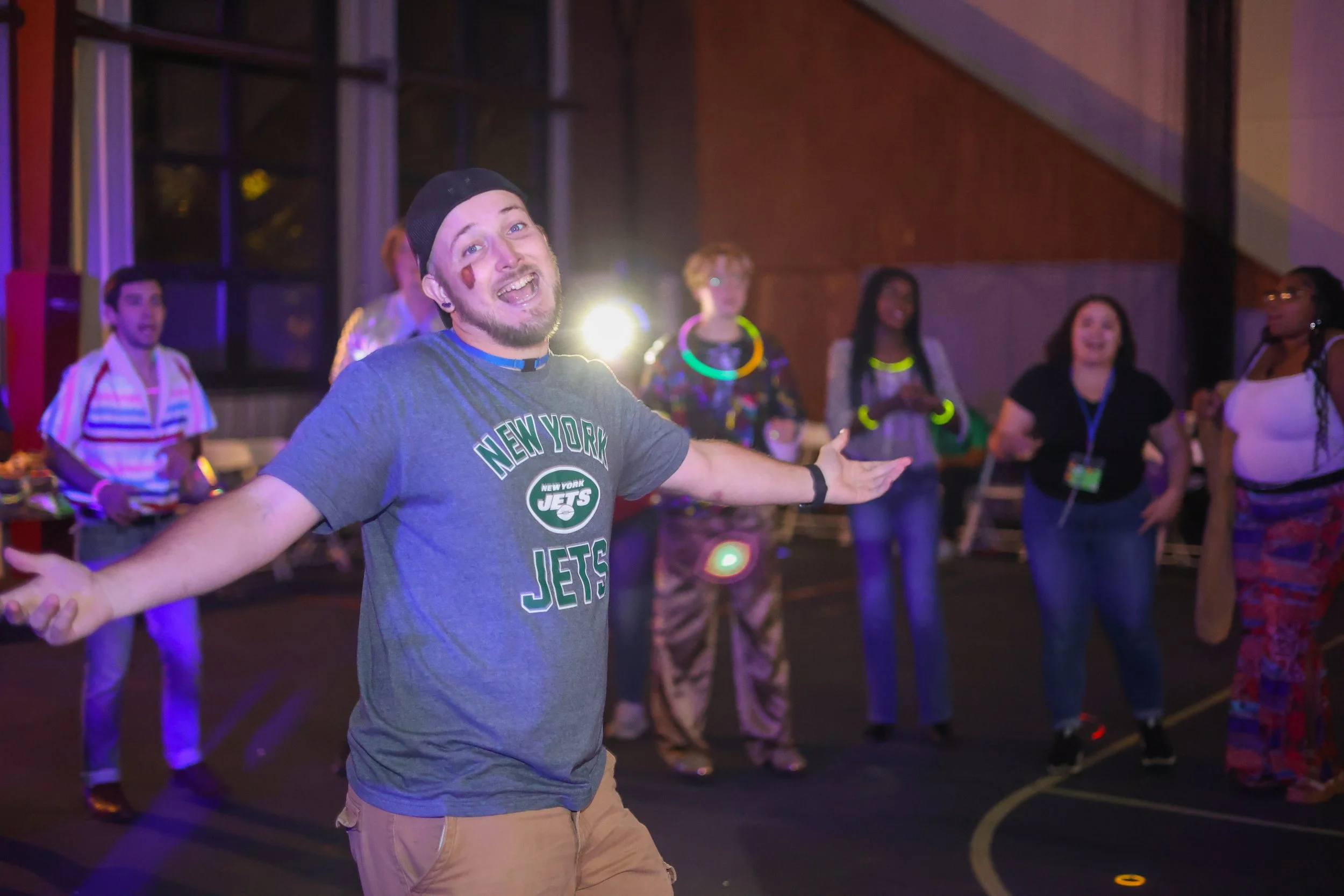 A man in a New York Jets t-shirt is smiling with his arms outstretched, dancing at a summer camp dance, with a group of campers cheering him on.