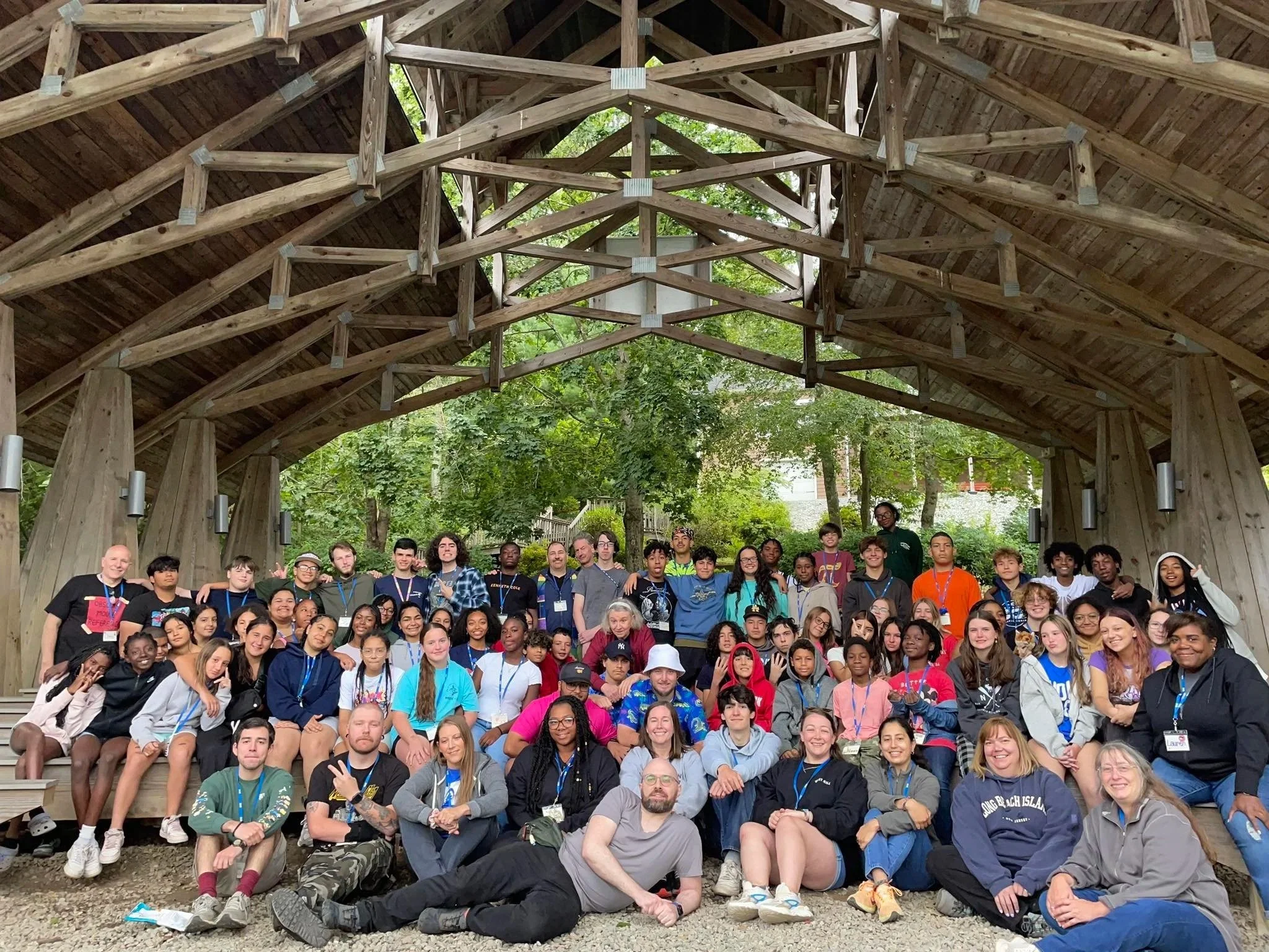 A large group of diverse young people and adults gathered together on a wooden stage outdoors under a wooden arch structure with trees in the background.