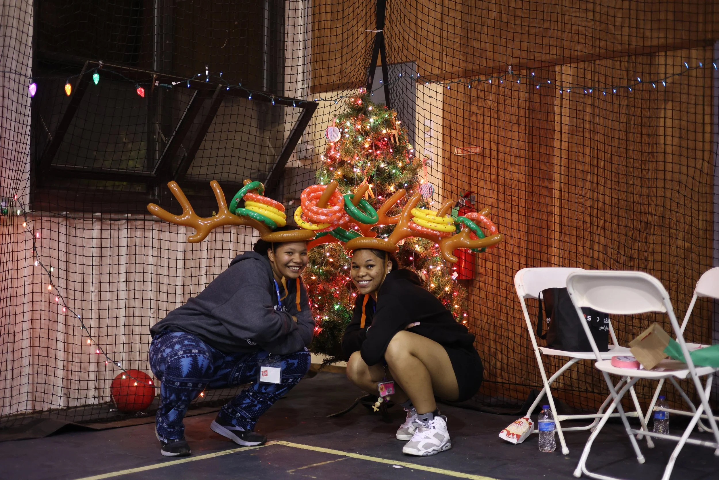 Two campers wearing reindeer antler headbands with balloon decorations, squatting in front of a decorated Christmas tree with lights, ornaments, and a star on top inside a venue with pink walls and netting.