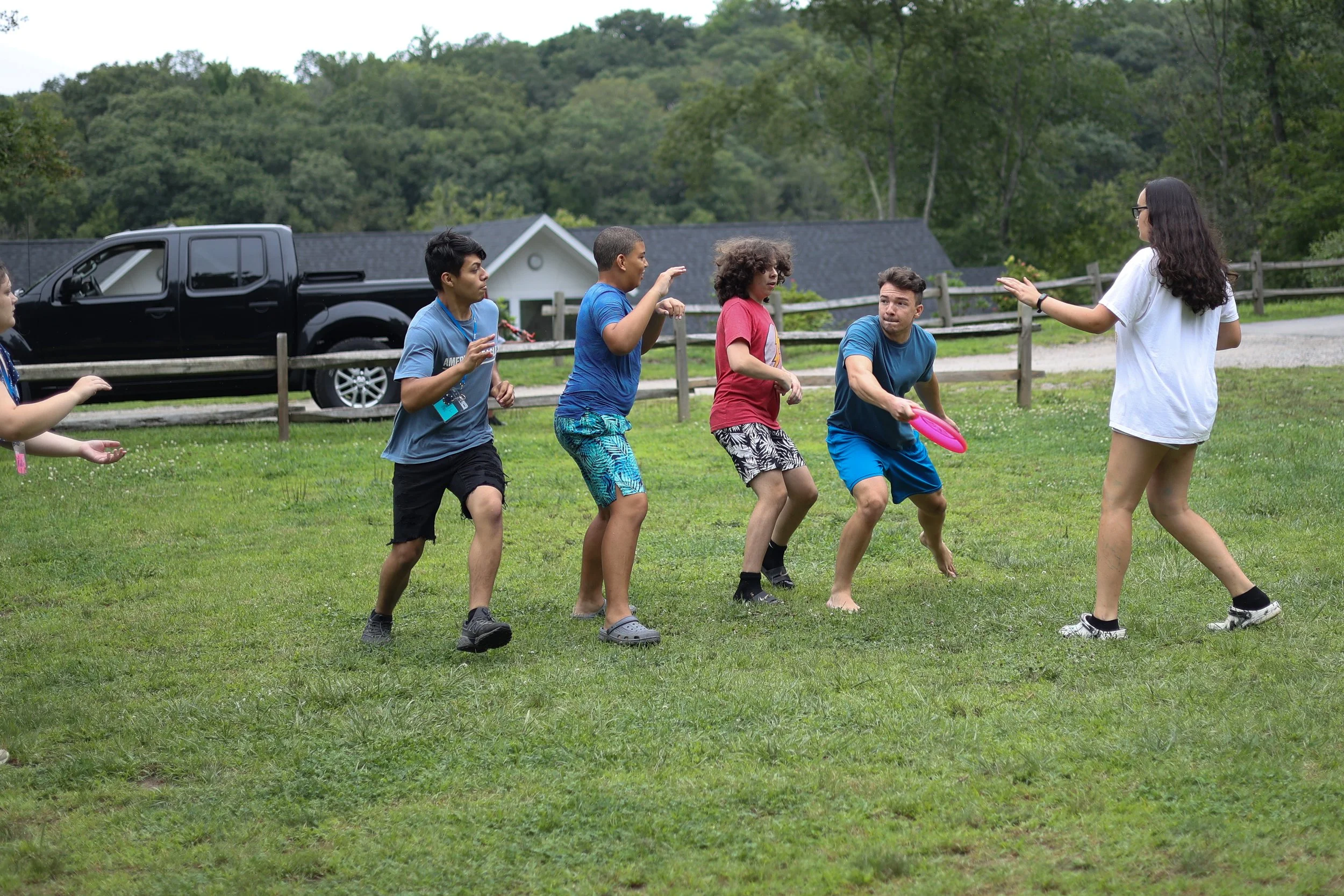 A group of five children playing frisbee on a grassy field with a woman supervising. They are outdoors with trees and a black truck in the background.