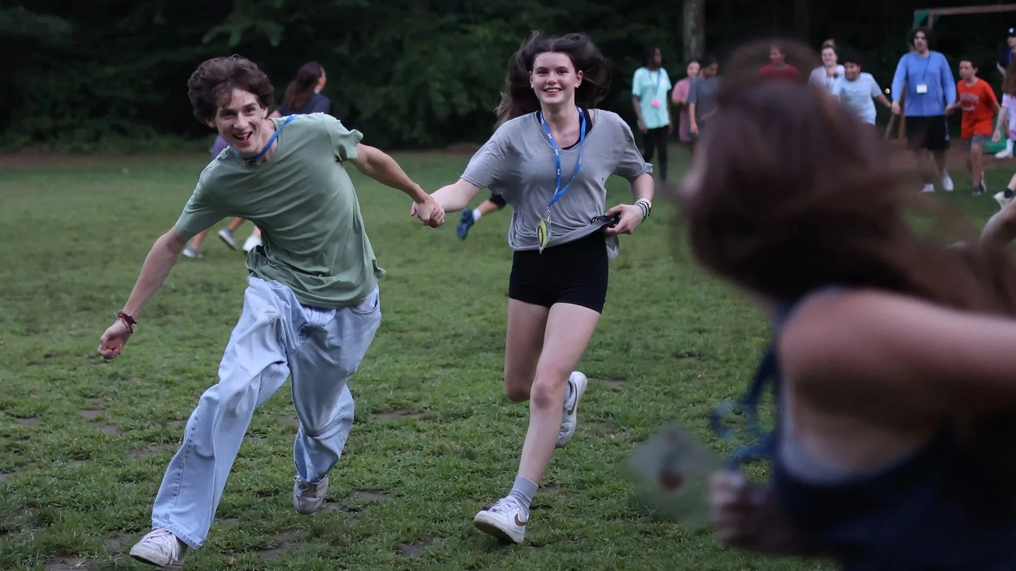 A group of teenagers running and playing outdoors on a grassy field during daytime, some holding hands and smiling.