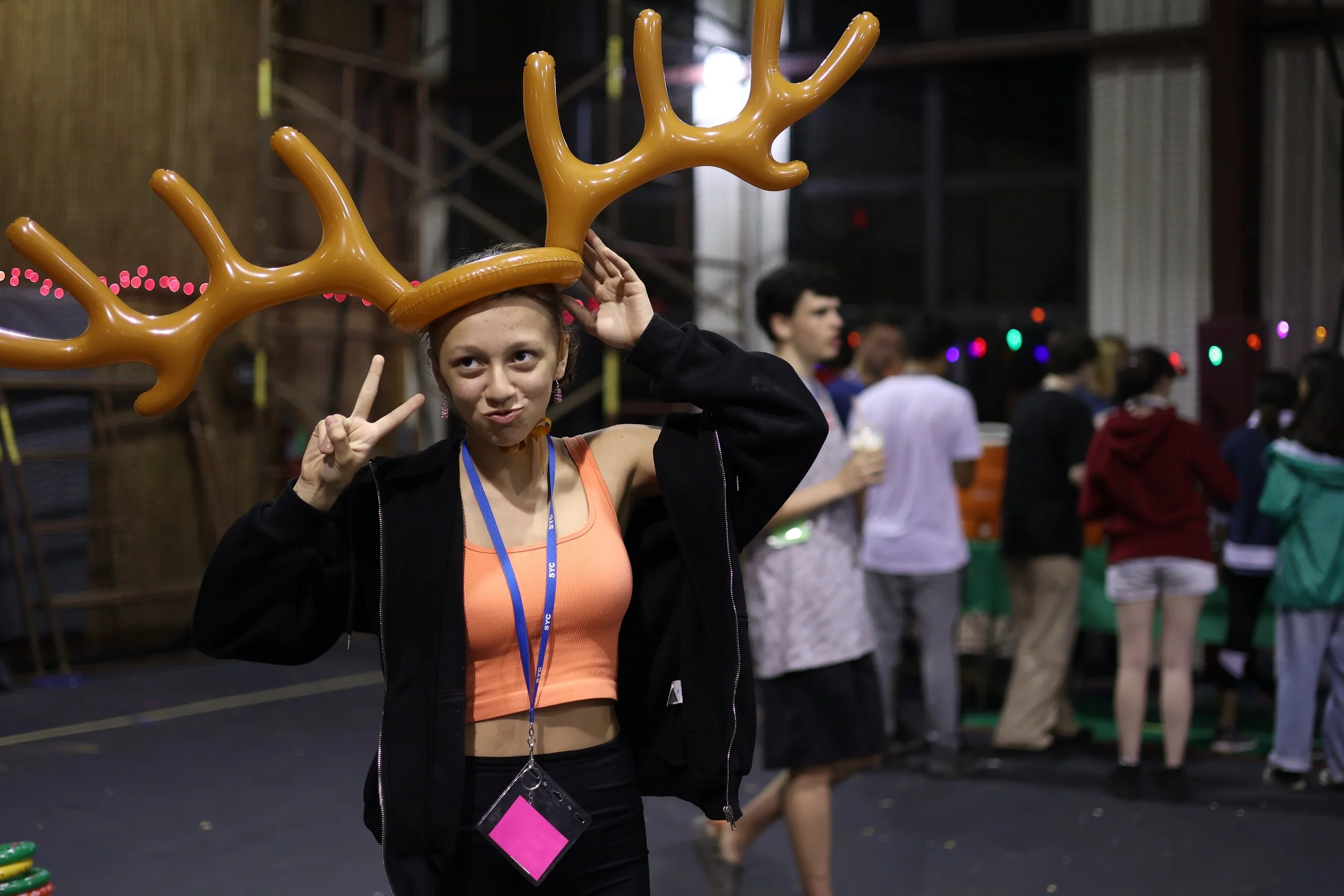 A camper wearing a reindeer antler headband making a peace sign at a festive event with people in line or socializing in the background.