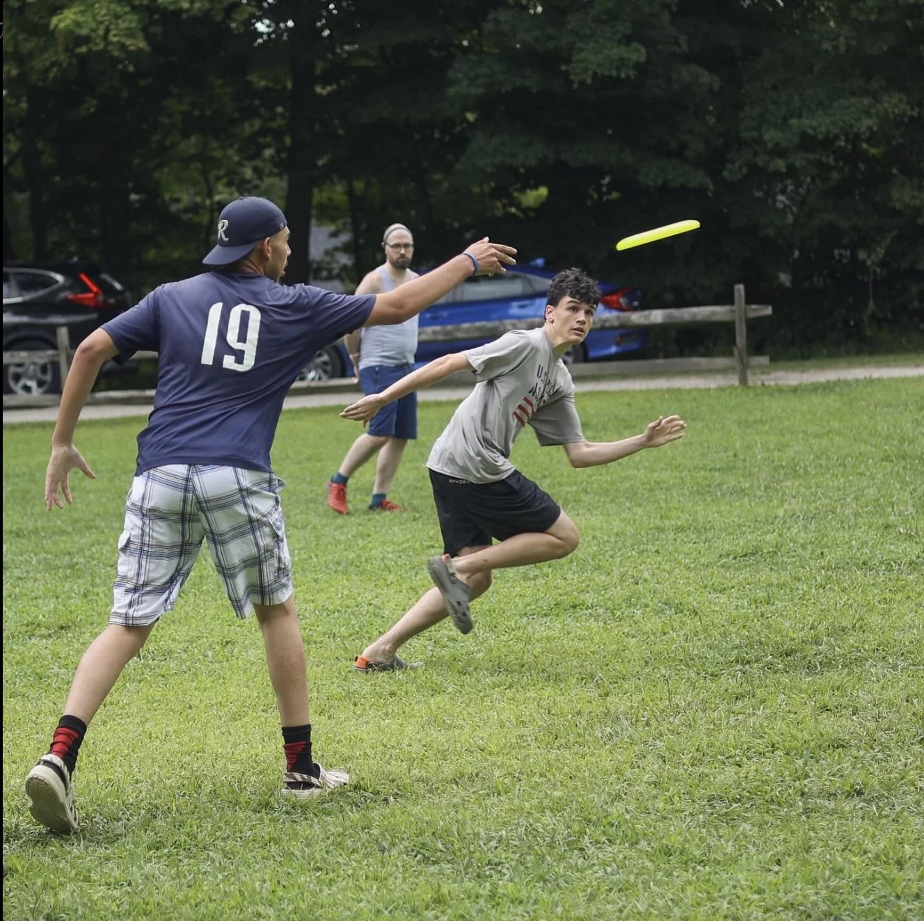 Three boys playing frisbee on a grassy field, with two parked cars and trees in the background.