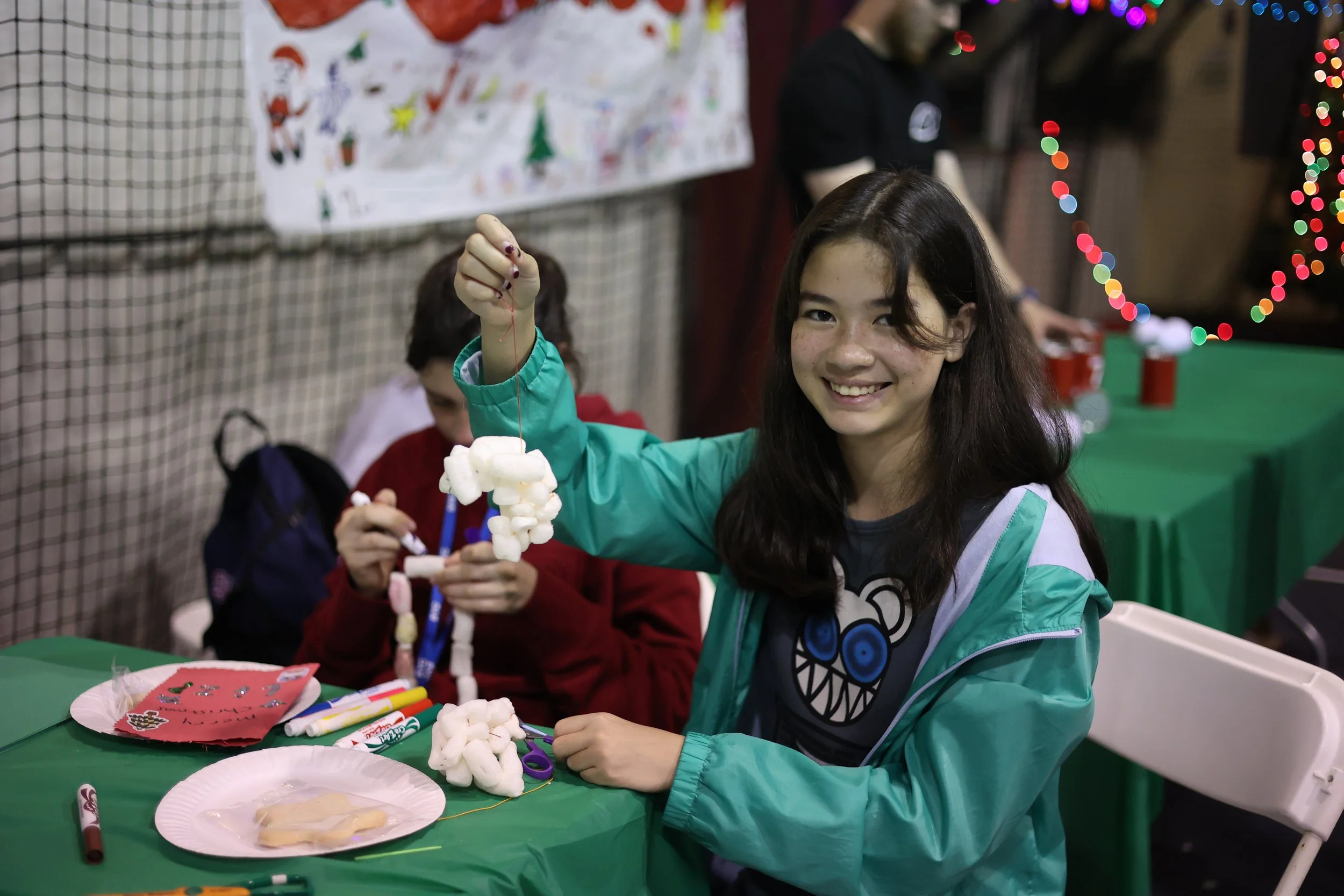 A girl with long dark hair wearing a teal jacket, smiling and holding up a handmade snowflake craft made with marshmallows, with other children and holiday decorations visible in the background.