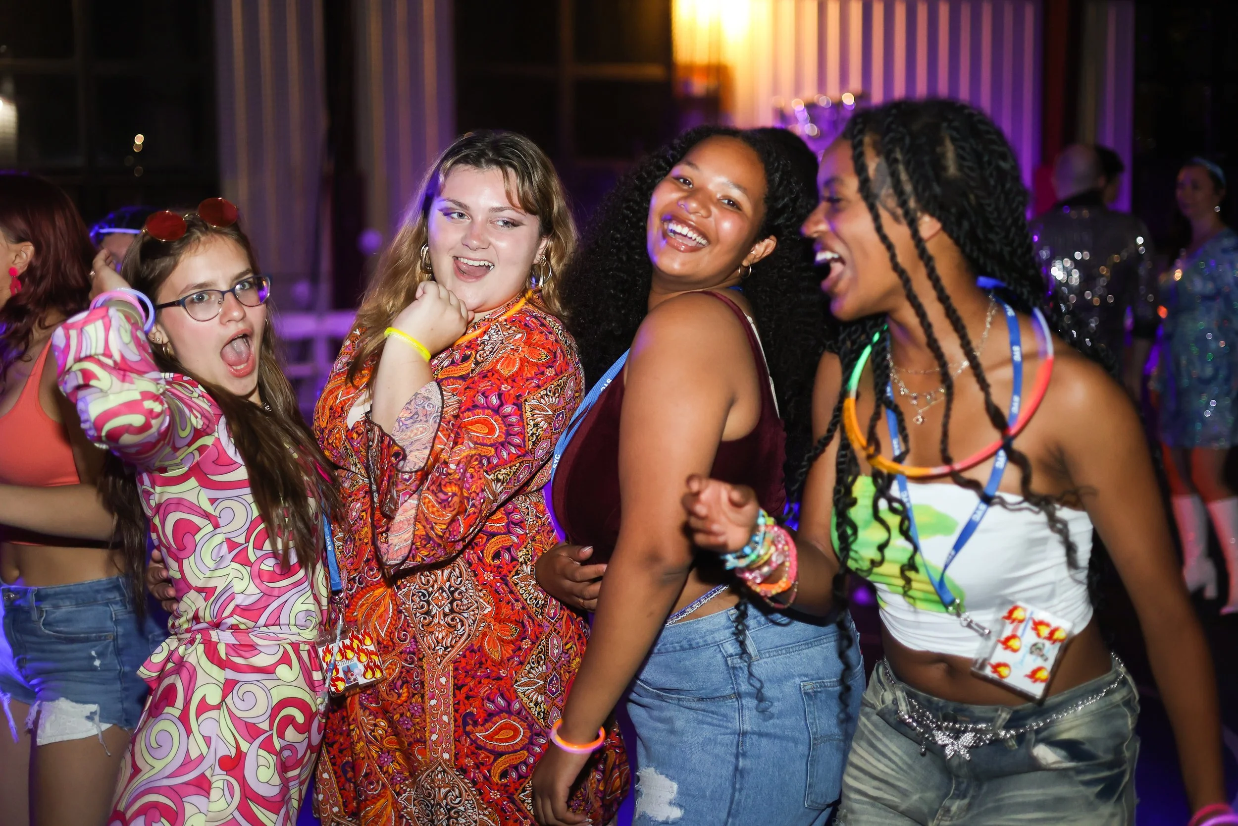 Four campers enjoying a summer camp dance, talking and laughing, with colorful lighting and other people in the background.