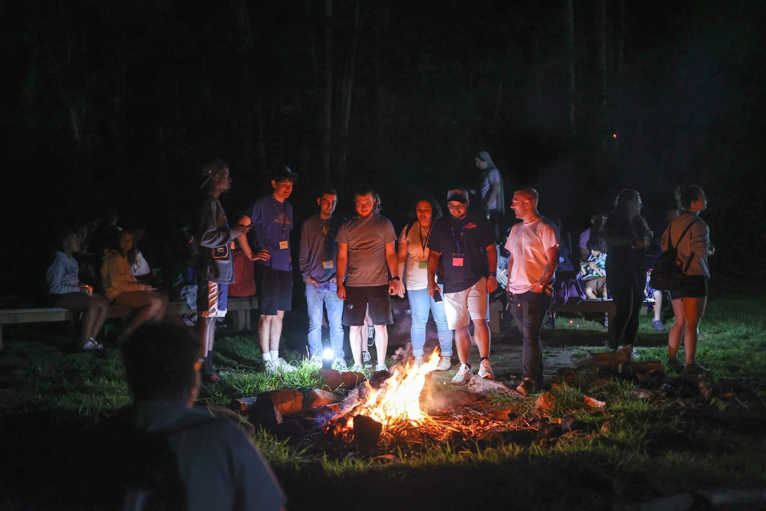 Group of people gathered around a campfire at night in an outdoor setting, some sitting on benches and others standing.