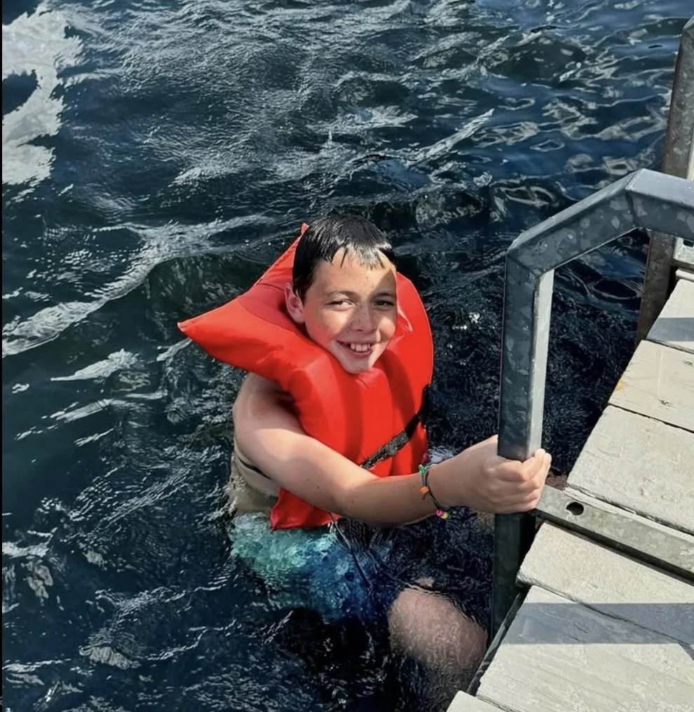 A smiling boy wearing a red life jacket climbing up a metal ladder into a boat, with water surrounding him.