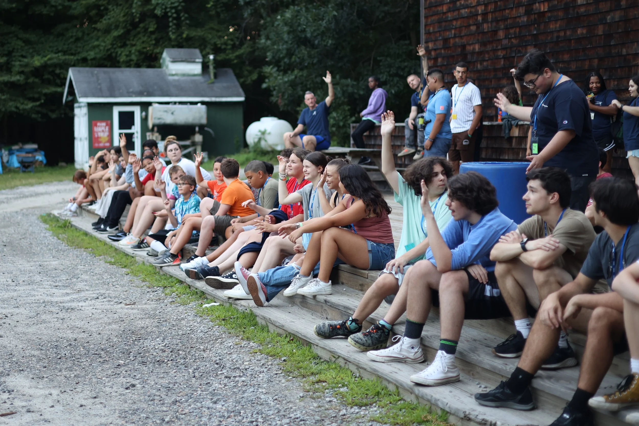 A group of children and teenagers sitting on wooden steps outdoors, with some raising their hands, during daytime.