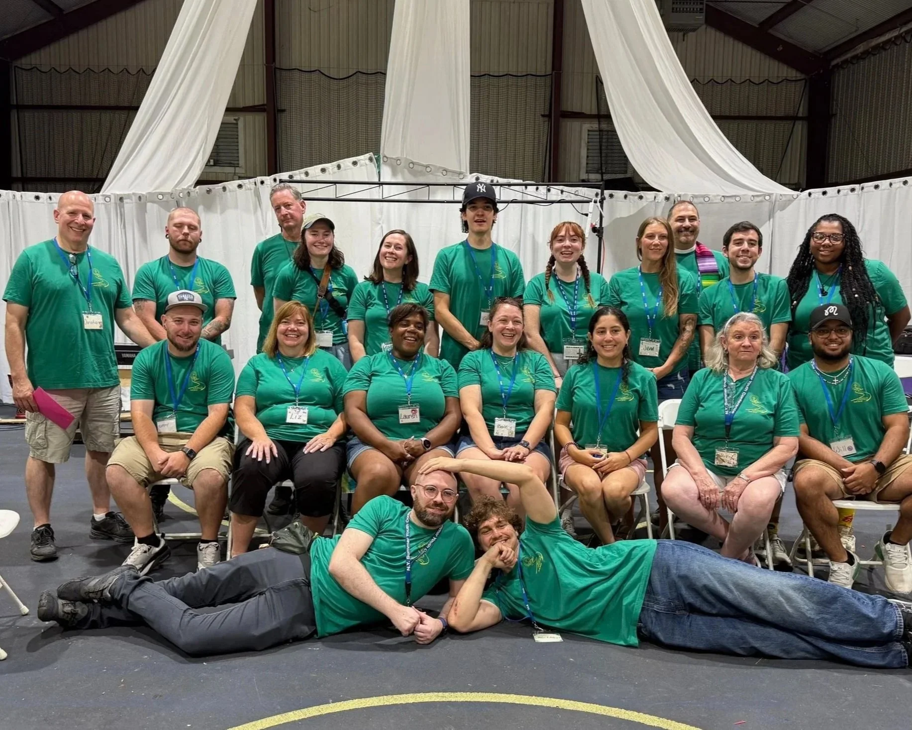 Group of people wearing matching green t-shirts posing for a photo indoors, some sitting on chairs, some standing, with a curtain backdrop.