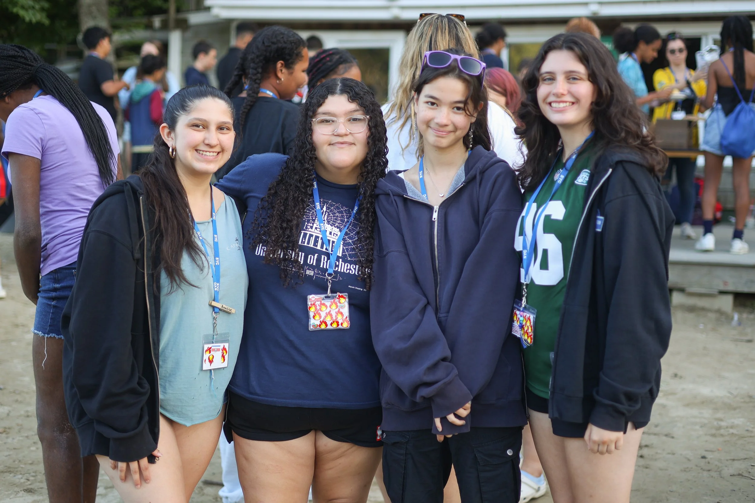 Four young women standing together outside at SYC, smiling at the camera, with other people in the background.