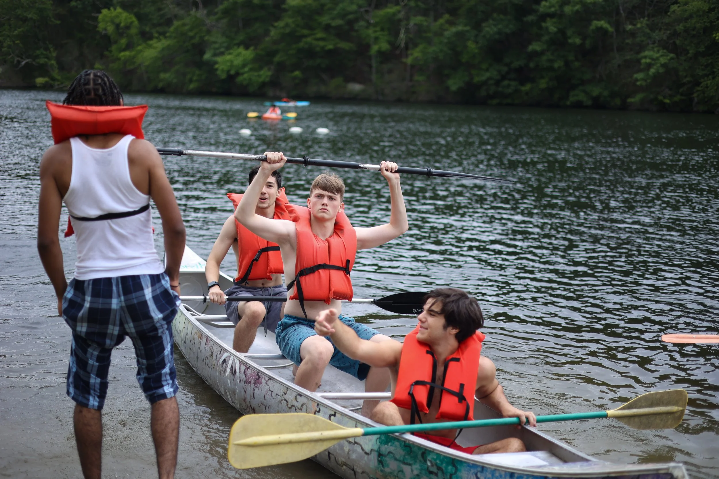 A group of young people in life jackets preparing to canoe on a lake surrounded by trees, with one person standing on the shore.
