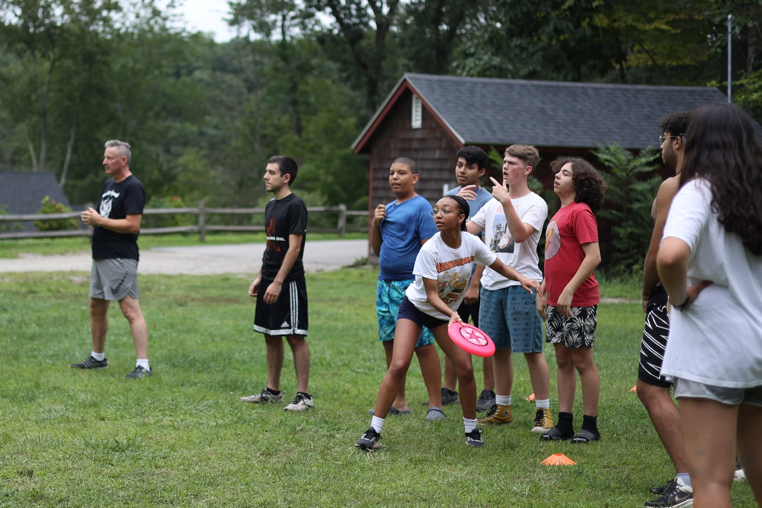 Group of children and adults standing on a grassy field, some holding frisbees, participating in an Ultimate Frisbee near a wooden building and trees.