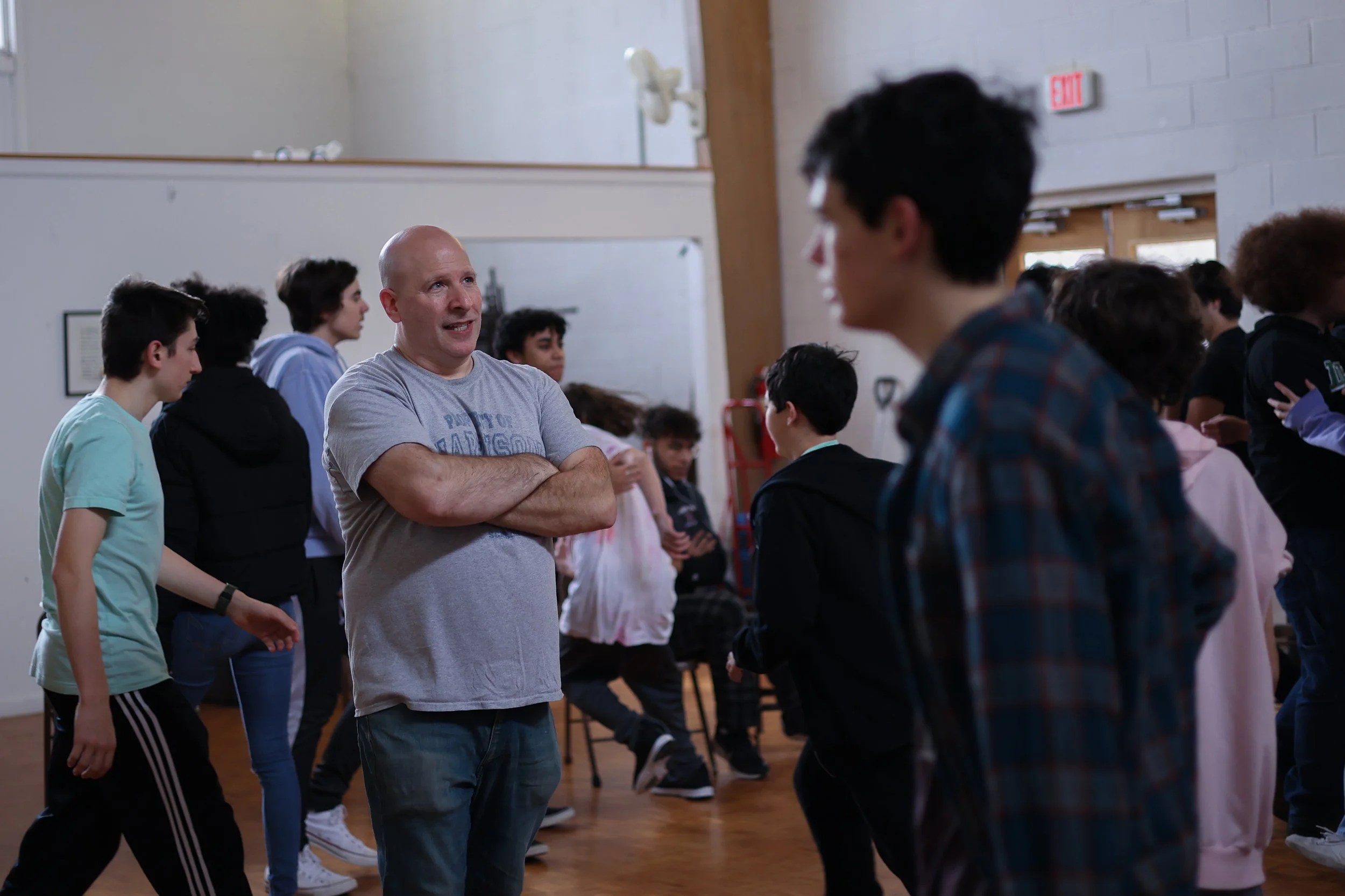 A group of teenagers in a gymnasium talking with an adult man with a bald head, wearing a grey t-shirt and jeans, who is smiling and has his arms crossed.