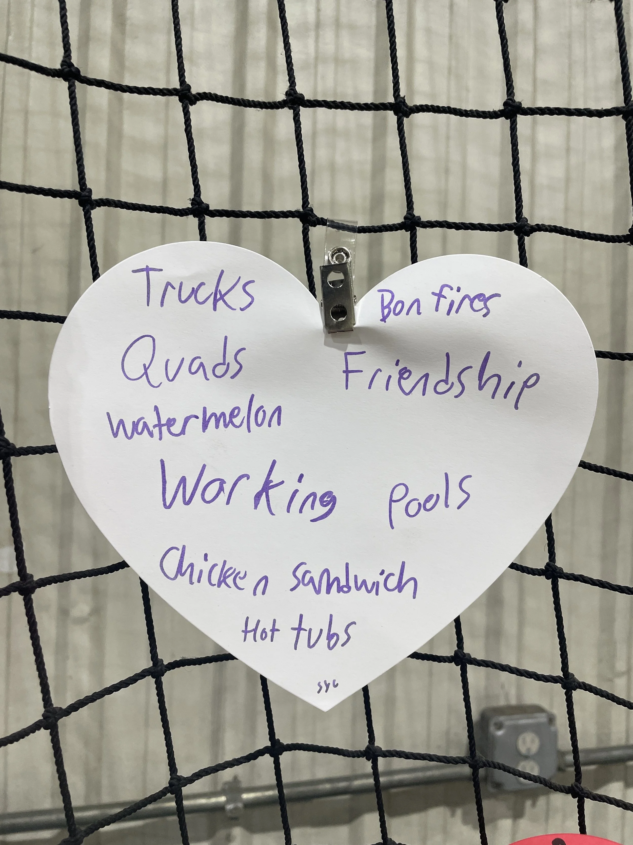 Heart-shaped note with handwritten list of items including trucks, quads, watermelon, working pools, chicken sandwich, hot tubs, bonfire, friendship, and salsa, hanging on a black wire fence.