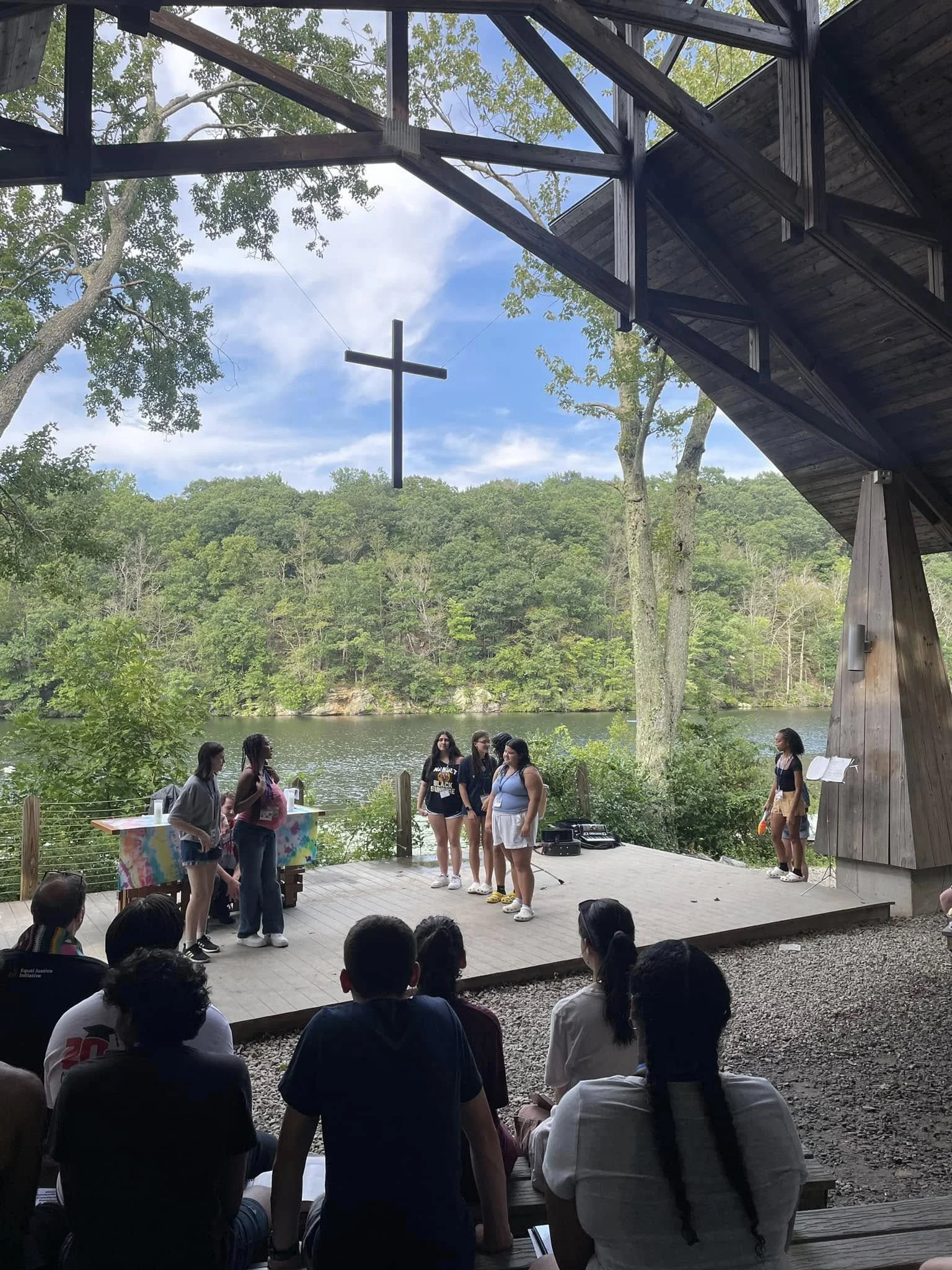 People performing on an outdoor stage near water with a hanging cross overhead and forest in the background.
