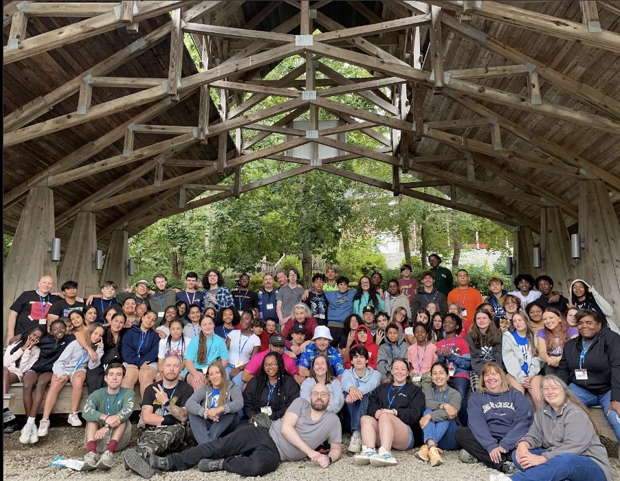 Group of diverse young people and adults gathered outdoors under a large wooden pavilion with a natural setting, smiling for a group photo.