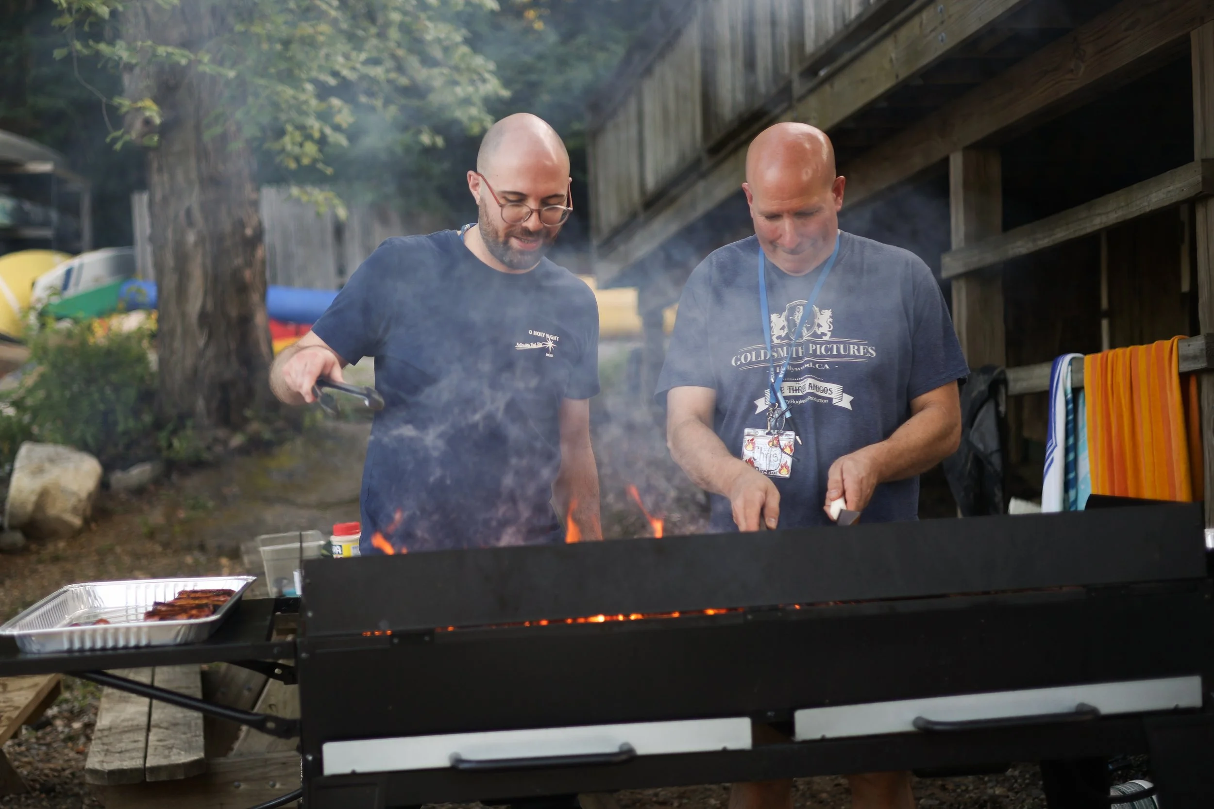 Two men grilling food outdoors, one using tongs to turn meat on a barbecue grill, with smoke rising, and a gravel area and wooden fence in the background.