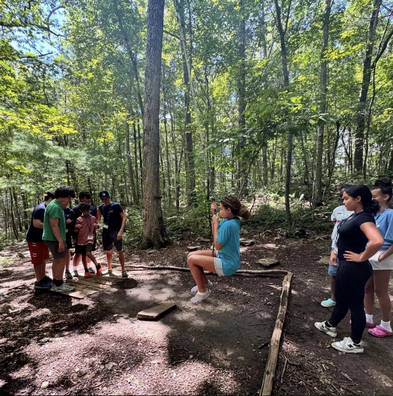 Teen girl sitting on a wooden log swing while a group of children and teenagers gather around in a forested area.