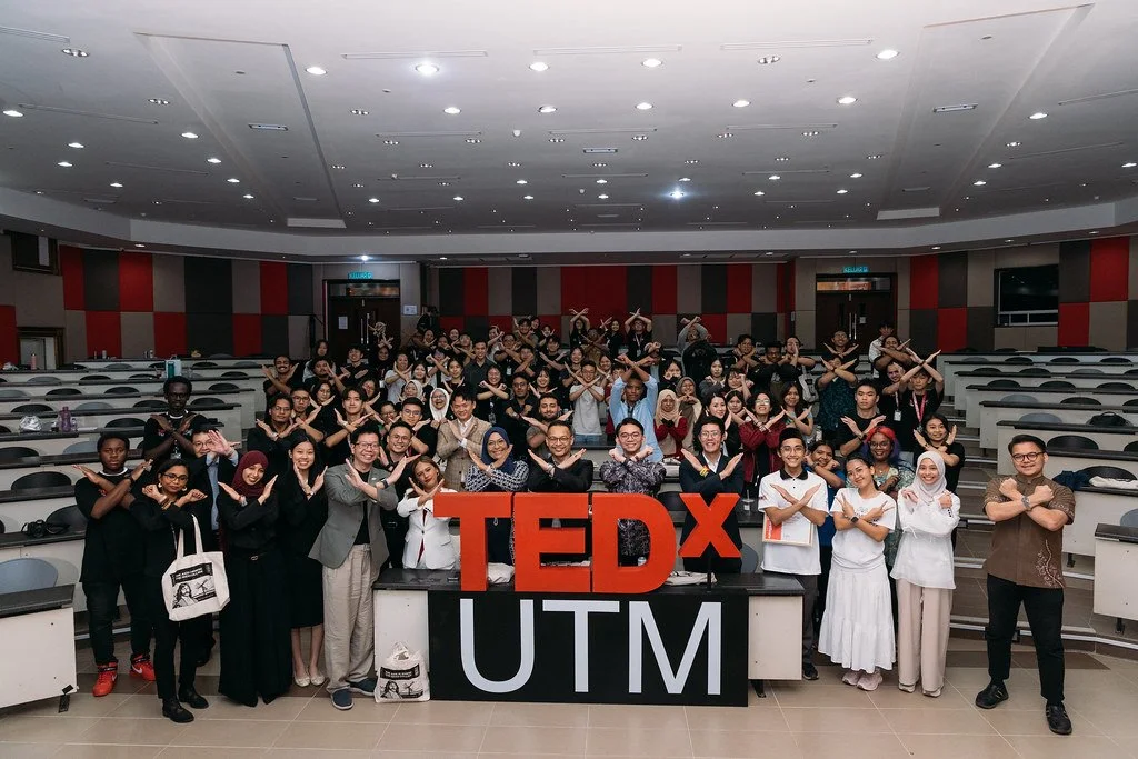 Dr-Khor-Kang-Xiang with group of diverse people participating in a TEDx event at UTM, standing in a lecture hall with a large red TEDx sign and crossing their arms in an X shape.