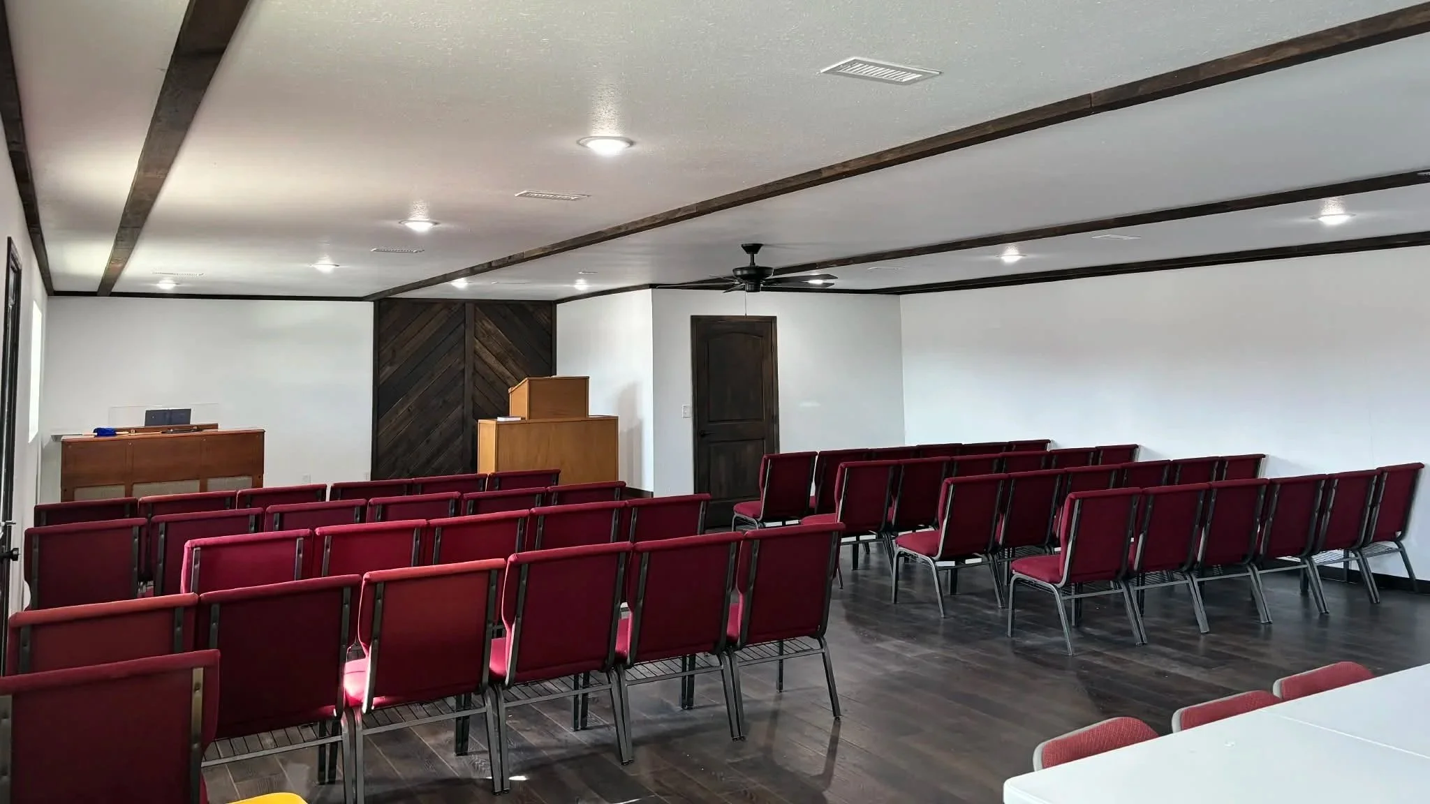 Empty sanctuary set up for worship services with rows of red chairs facing a small wooden podium and a black lectern, white walls, a dark wood-paneled accent wall, and a ceiling with recessed lighting and a ceiling fan.