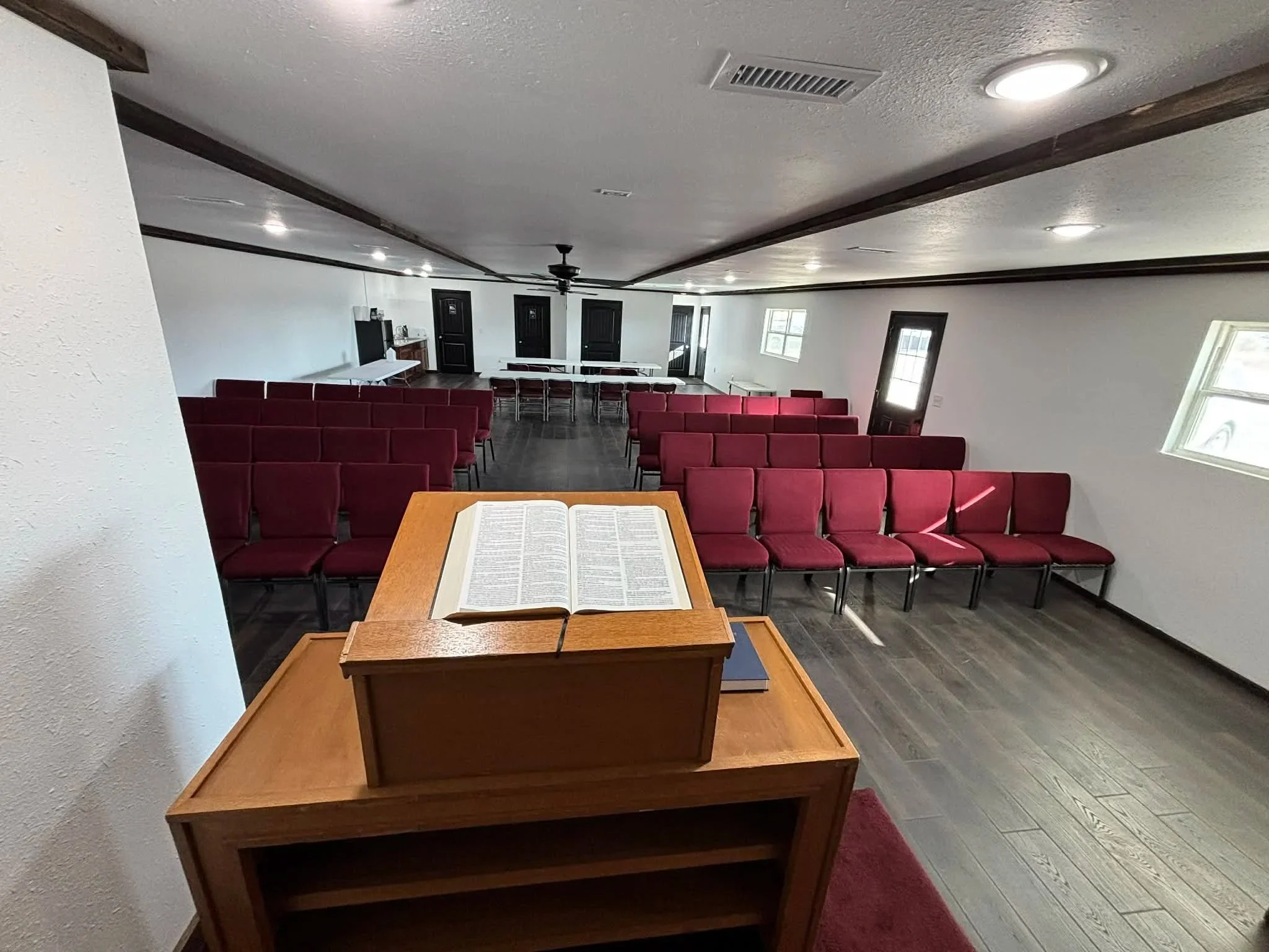 Interior of a small church with red cushioned chairs arranged in rows, a lectern with an open Bible at the front, white walls, windows on the sides, and doors at the back, with a ceiling fan and lights.