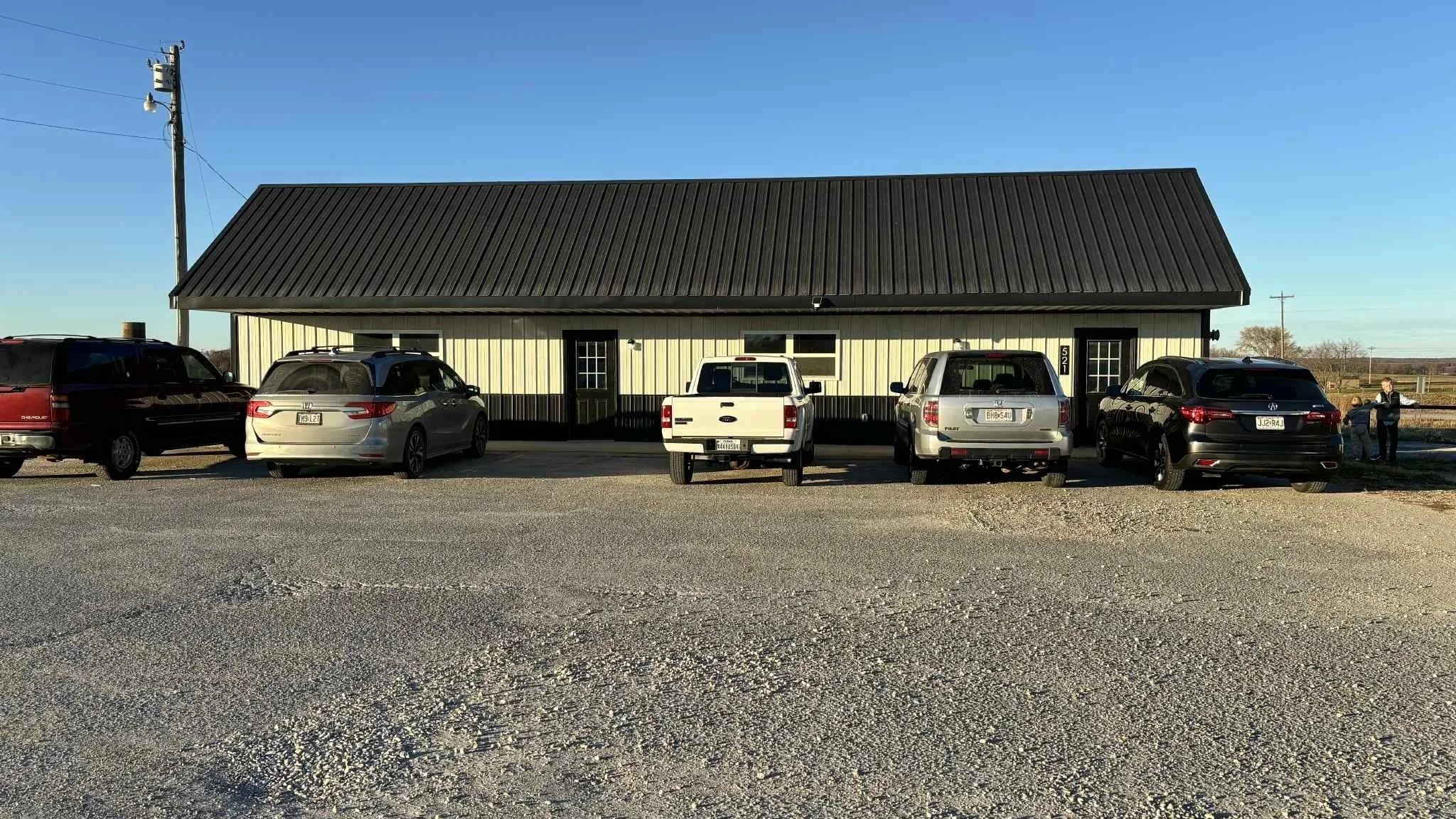 Parking lot in front of a building with a metal roof, five cars parked, and two people standing on the right side.