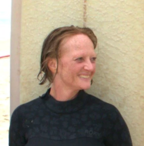 A woman with reddish-brown hair smiling and looking to the side, wearing a black top, standing in front of a textured beige wall.
