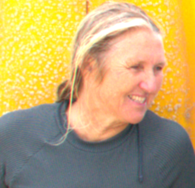 A woman with blonde hair smiling, wearing a dark gray shirt, standing in front of a yellow background.