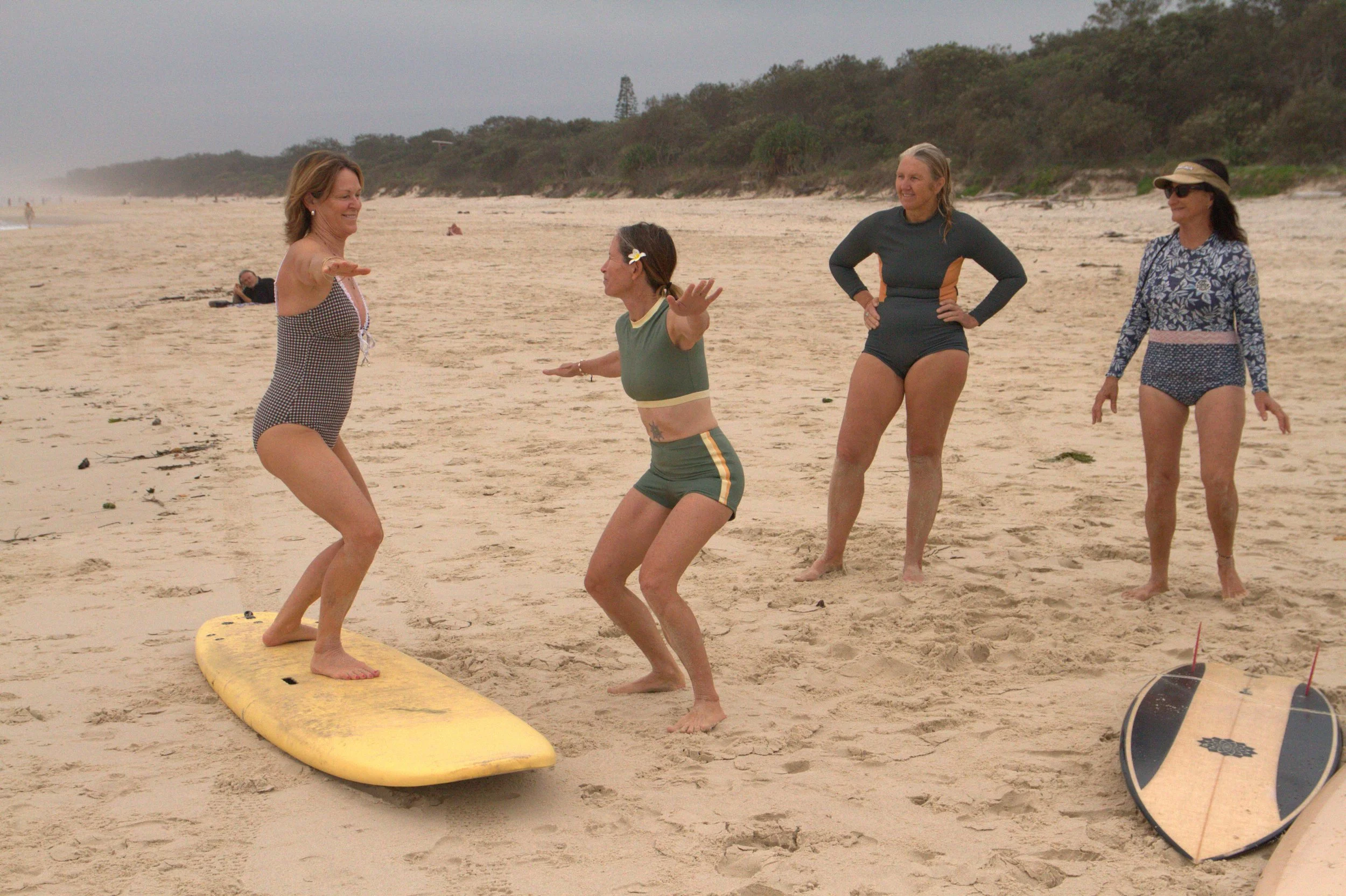 Four women on a sandy beach, one on a yellow surfboard, the others standing nearby, engaging in a conversation or laughing, with trees and a cloudy sky in the background.