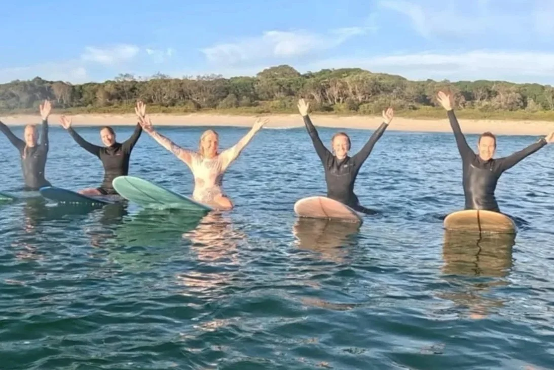 Five women in wetsuits on surfboards celebrating in the ocean with a coastal landscape behind them.
