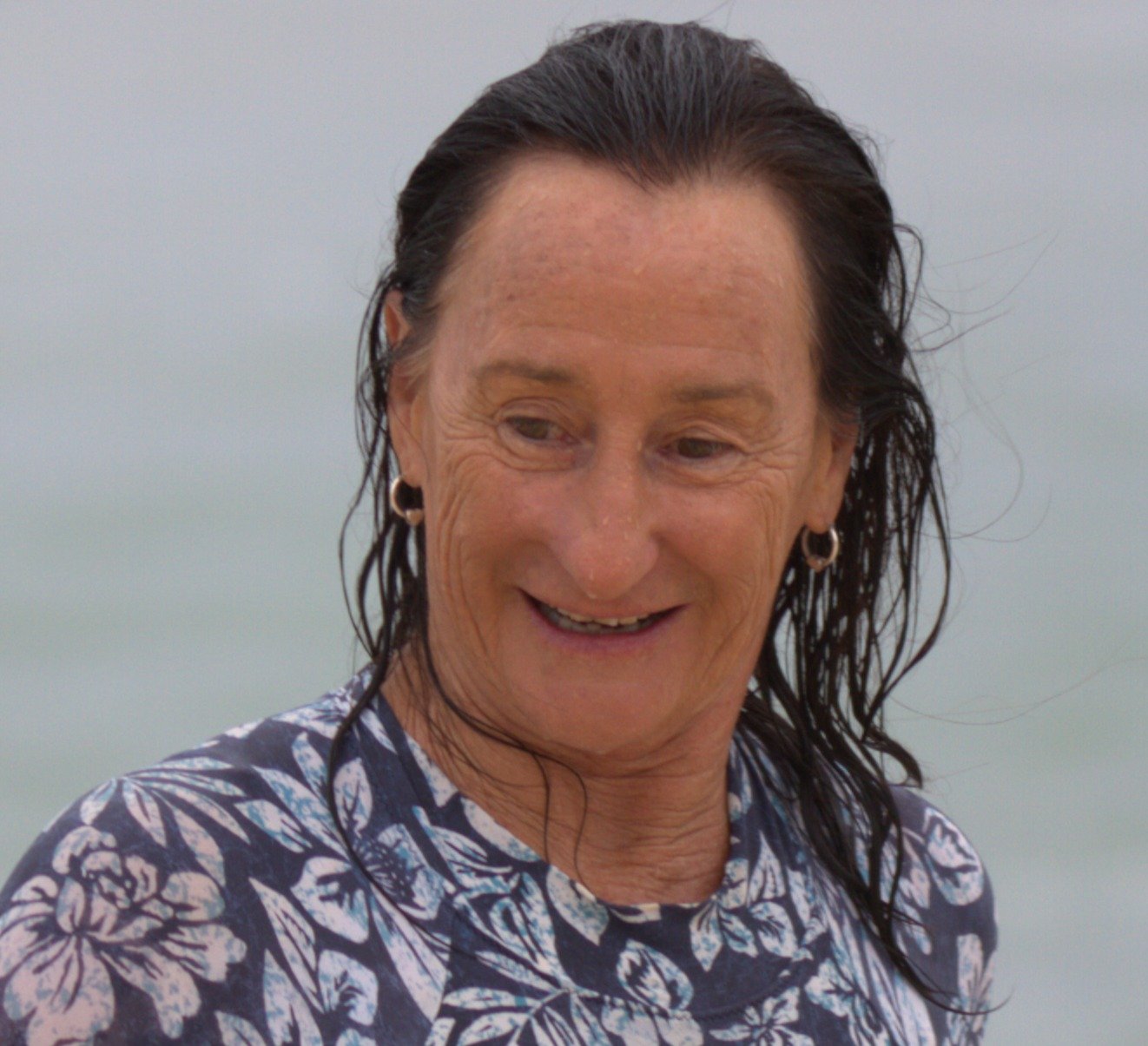 A smiling woman with wet, dark hair, wearing earrings and a patterned shirt, standing outdoors with a cloudy sky in the background.