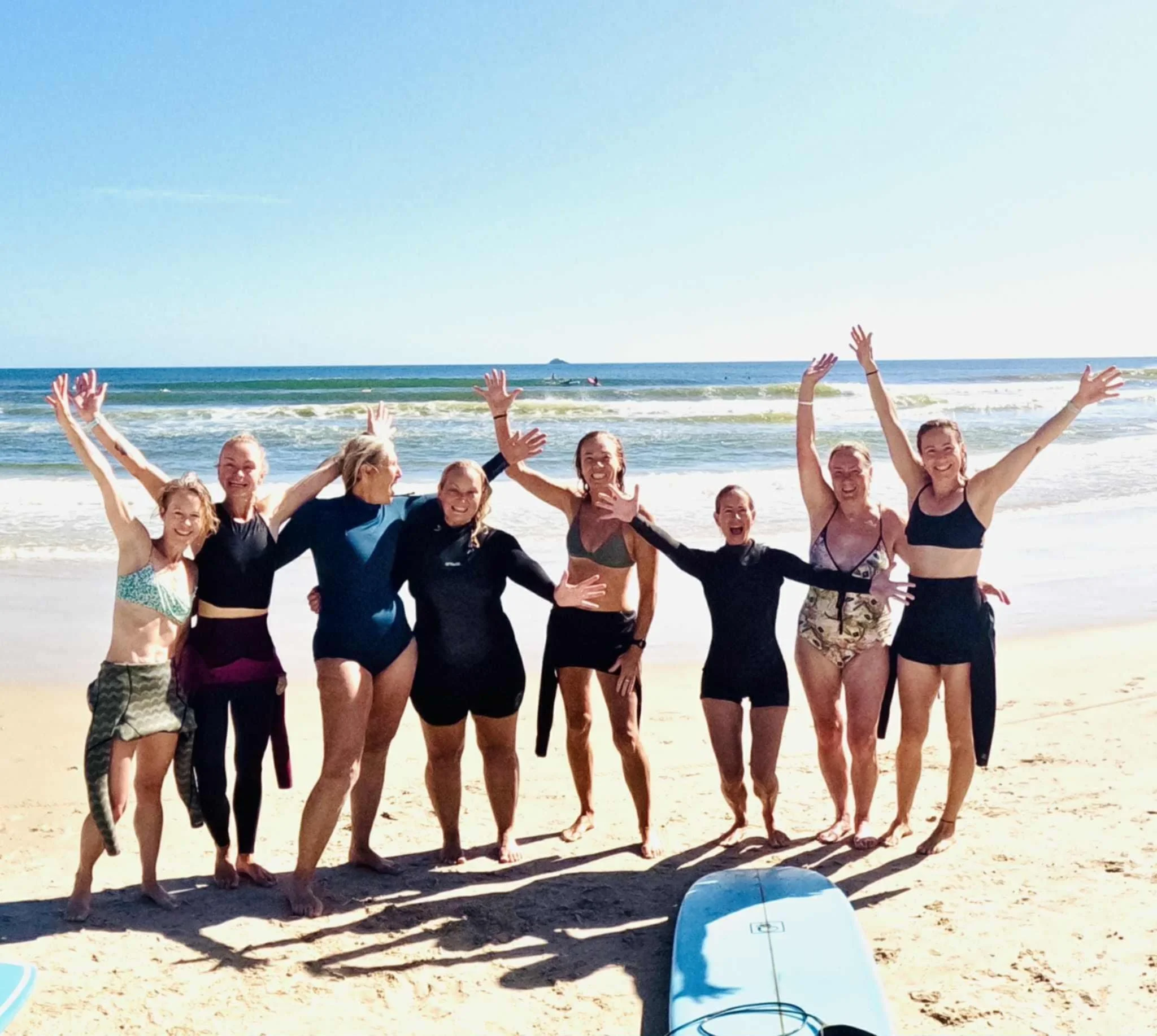 Group of women in swimsuits and wetsuits smiling and posing on the beach near the ocean with surfboard in foreground.