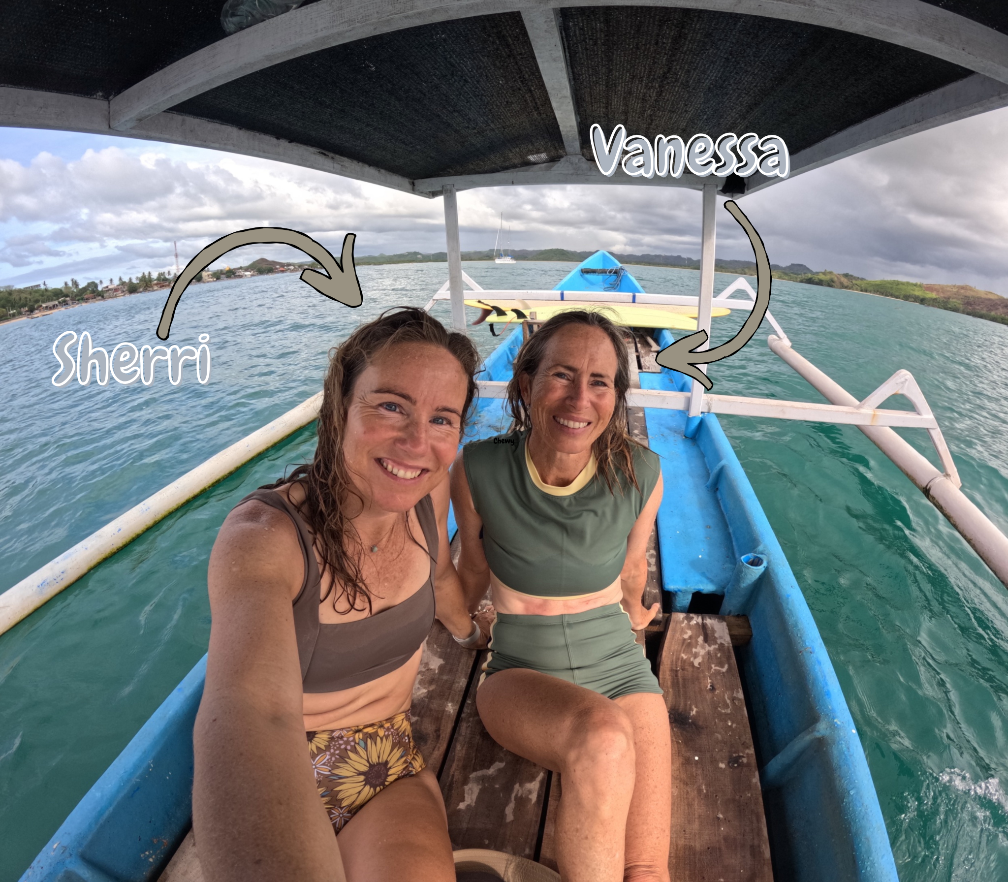 Two women, Sherri and Vanessa, sitting on a boat in the water with a shoreline and cloudy sky in the background.