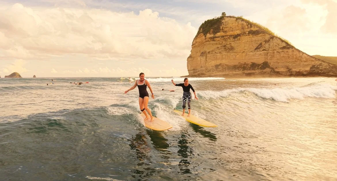 Two women surfing on small waves near a large coastal rock formation during sunset.