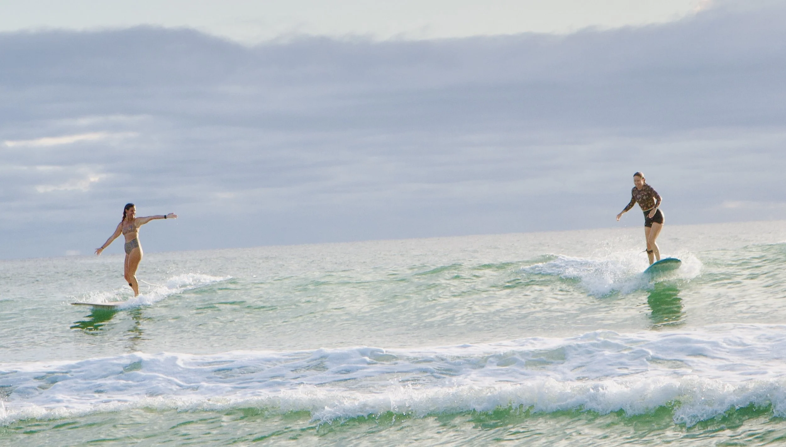Two women surfing on the ocean, one riding a wave and the other standing on her surfboard with arms outstretched, under a cloudy sky.
