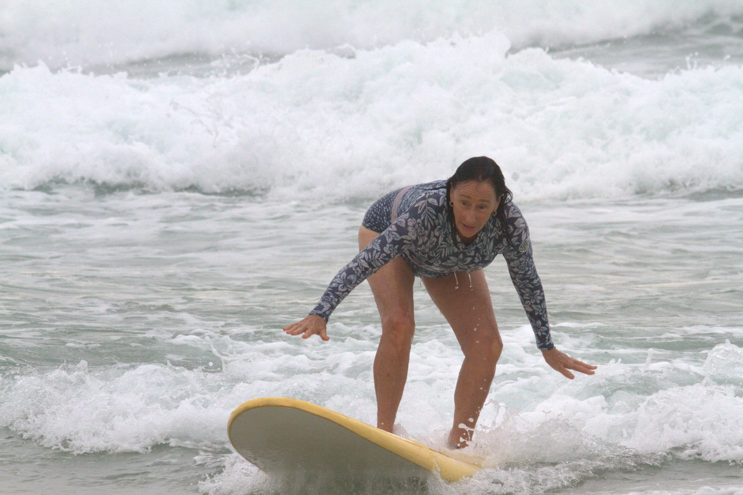 Woman in floral patterned long sleeve top and shorts surfing on a yellow surfboard in the ocean