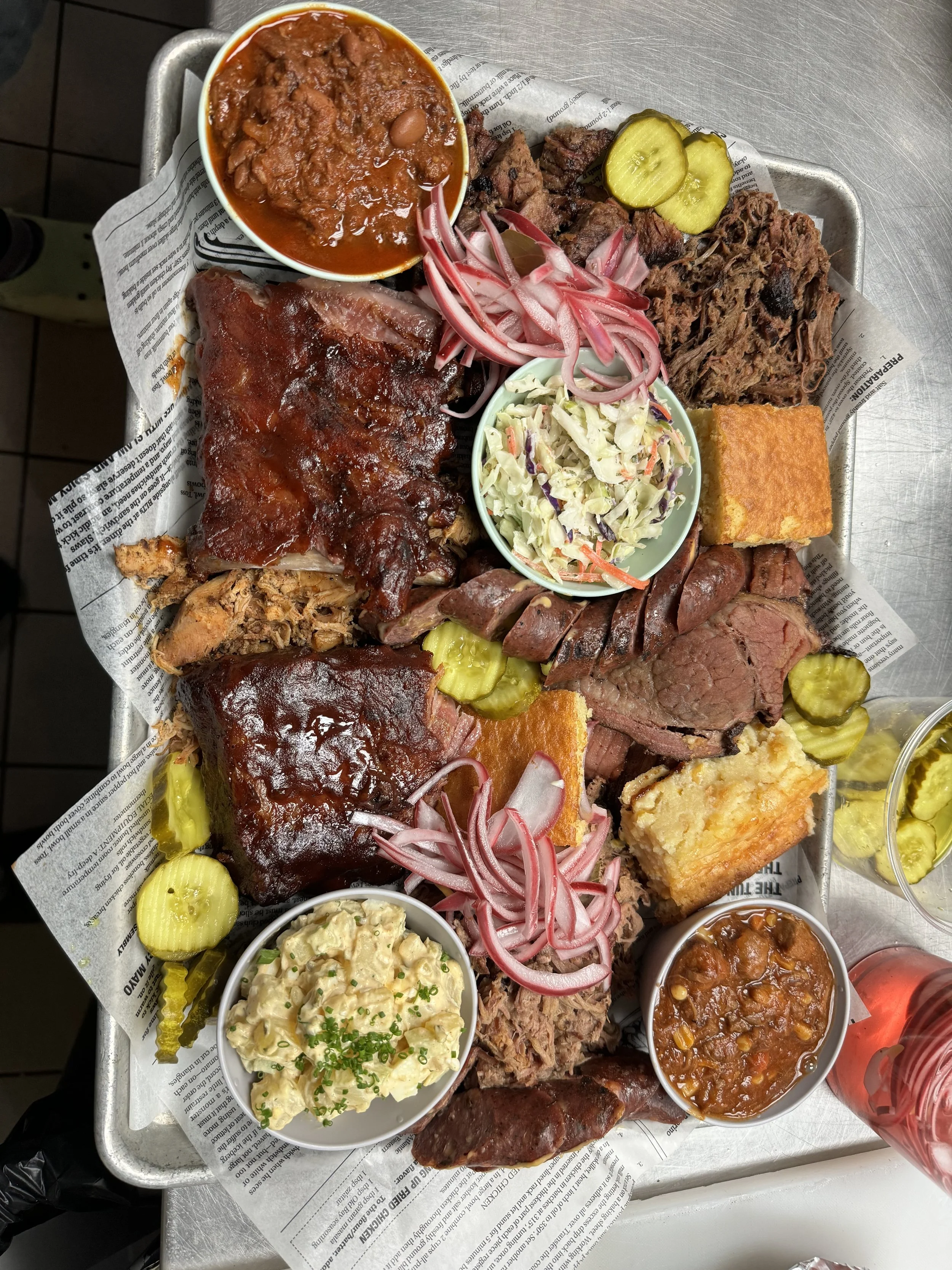 A tray of assorted barbecue meats, including ribs, brisket, sausage, and pulled pork, garnished with pickles, sliced onions, and served with sides like potato salad, coleslaw, baked beans, cornbread, and chili.