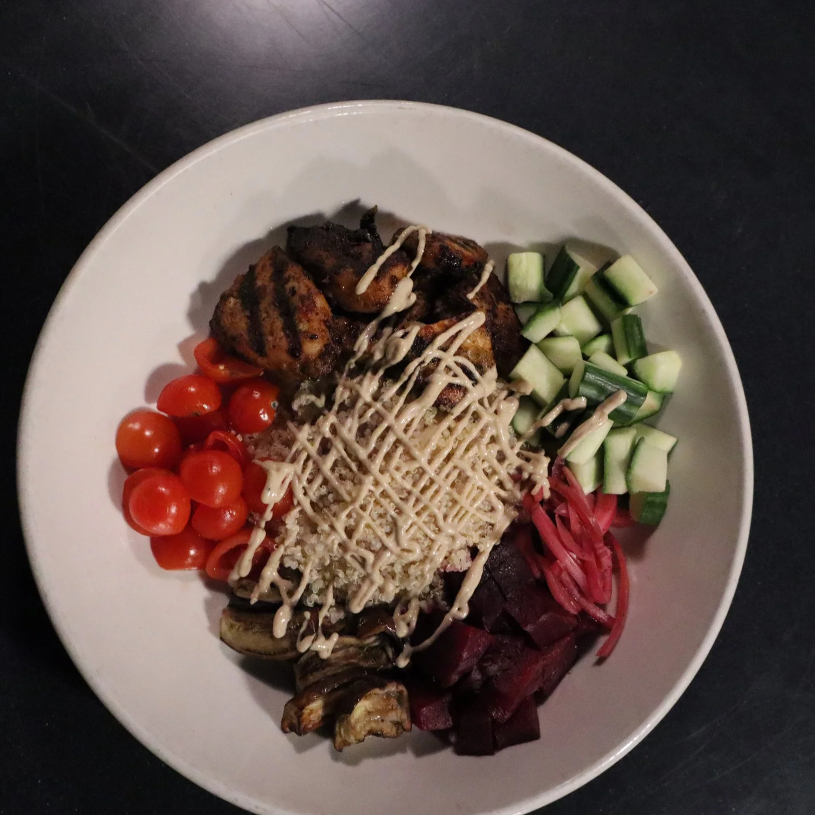 Plated dish with grilled chicken, cherry tomatoes, chopped cucumbers, shredded beet, eggplant, and a drizzle of tahina sauce on a white plate.