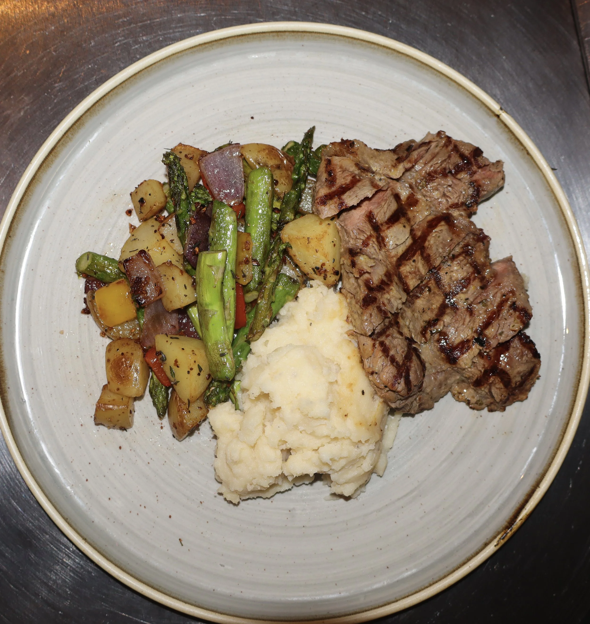 A plate with grilled steak, mashed potatoes, and a side of roasted vegetables including potatoes, asparagus, red onion, and red bell pepper.