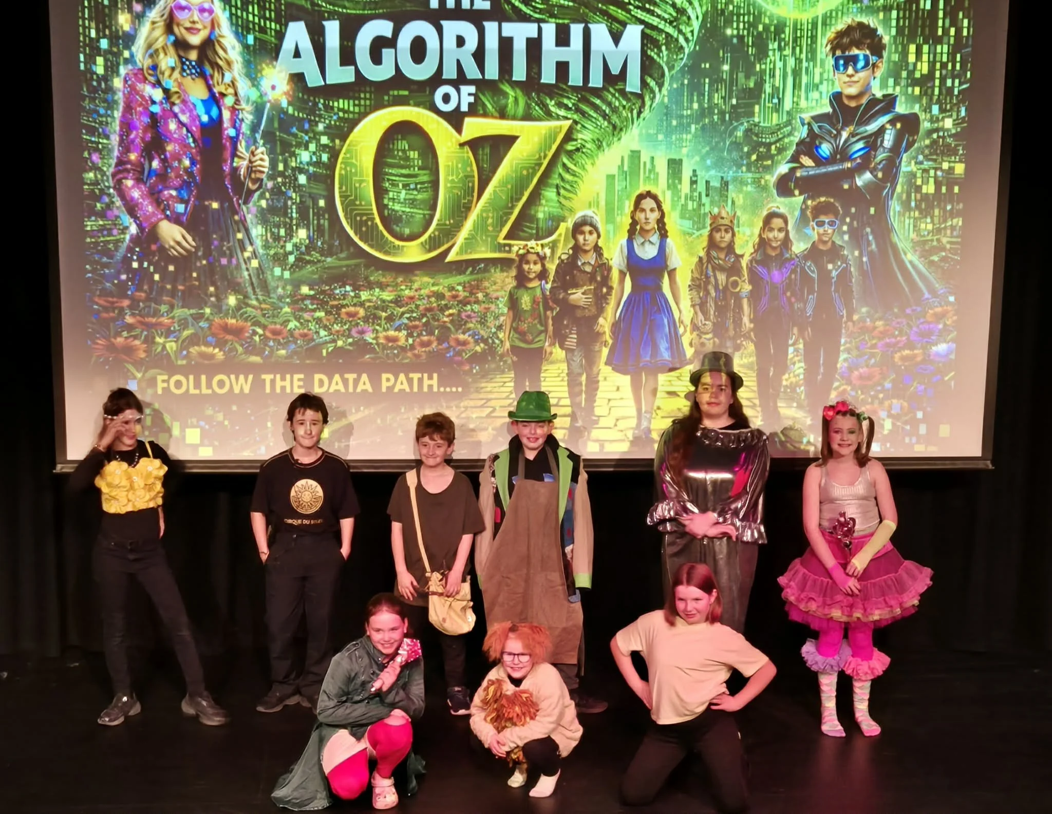 Children dressed in colorful costumes on stage in front of a vibrant backdrop with the text 'Algorithm of Oz' and children in various costumes.