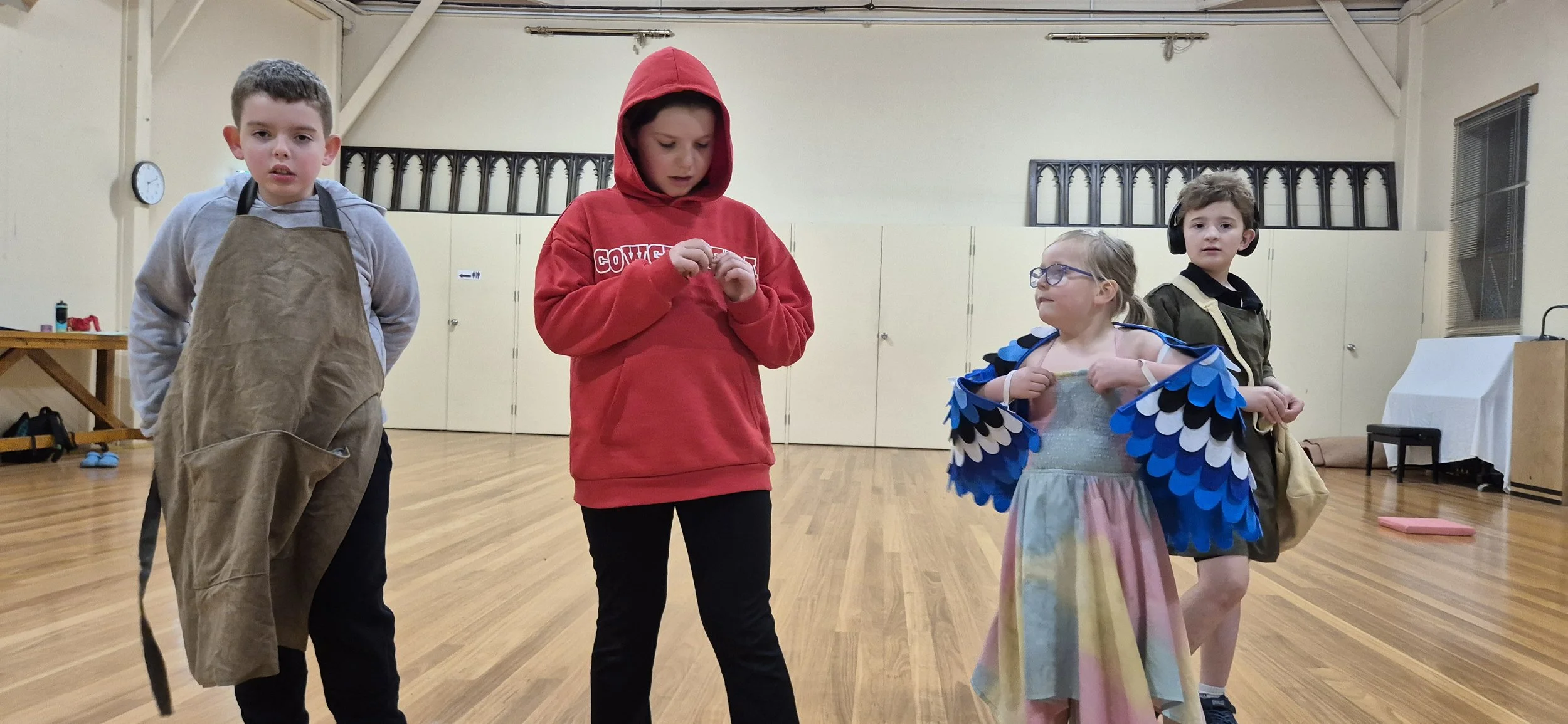 Four children standing in a room, one dressed as a bird, others in casual clothing, some with headphones, in a dance studio or classroom with wooden floors and a clock on the wall.