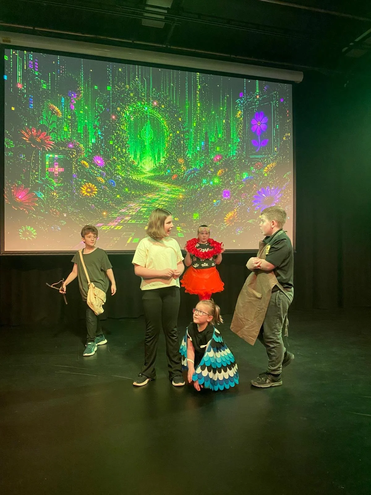A group of children performing on stage, with a bright digital fantasy background projected behind them, including flowers and neon colors.