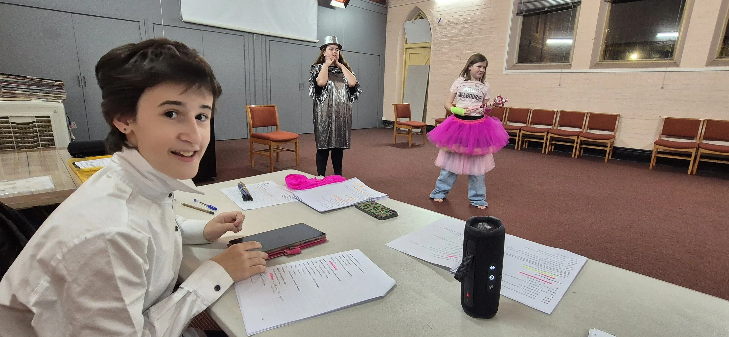 A boy with short dark hair smiling at the camera, seated at a table with papers, a notebook, and a phone. In the background, two girls are performing, one with a metallic dress and a silver hat, and the other wearing a pink tutu and holding a wand.