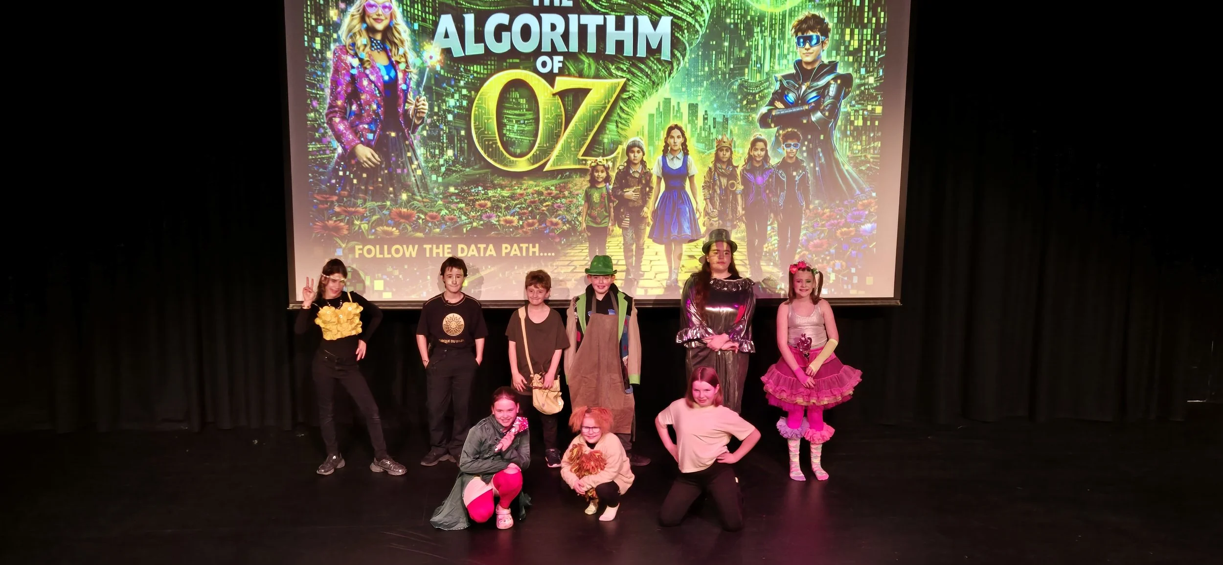 Group of children in costumes posing on stage in front of a large colorful digital poster for The Algorithm of Oz with characters from the Wizard of Oz, including a girl dressed as Dorothy and others in various costumes.