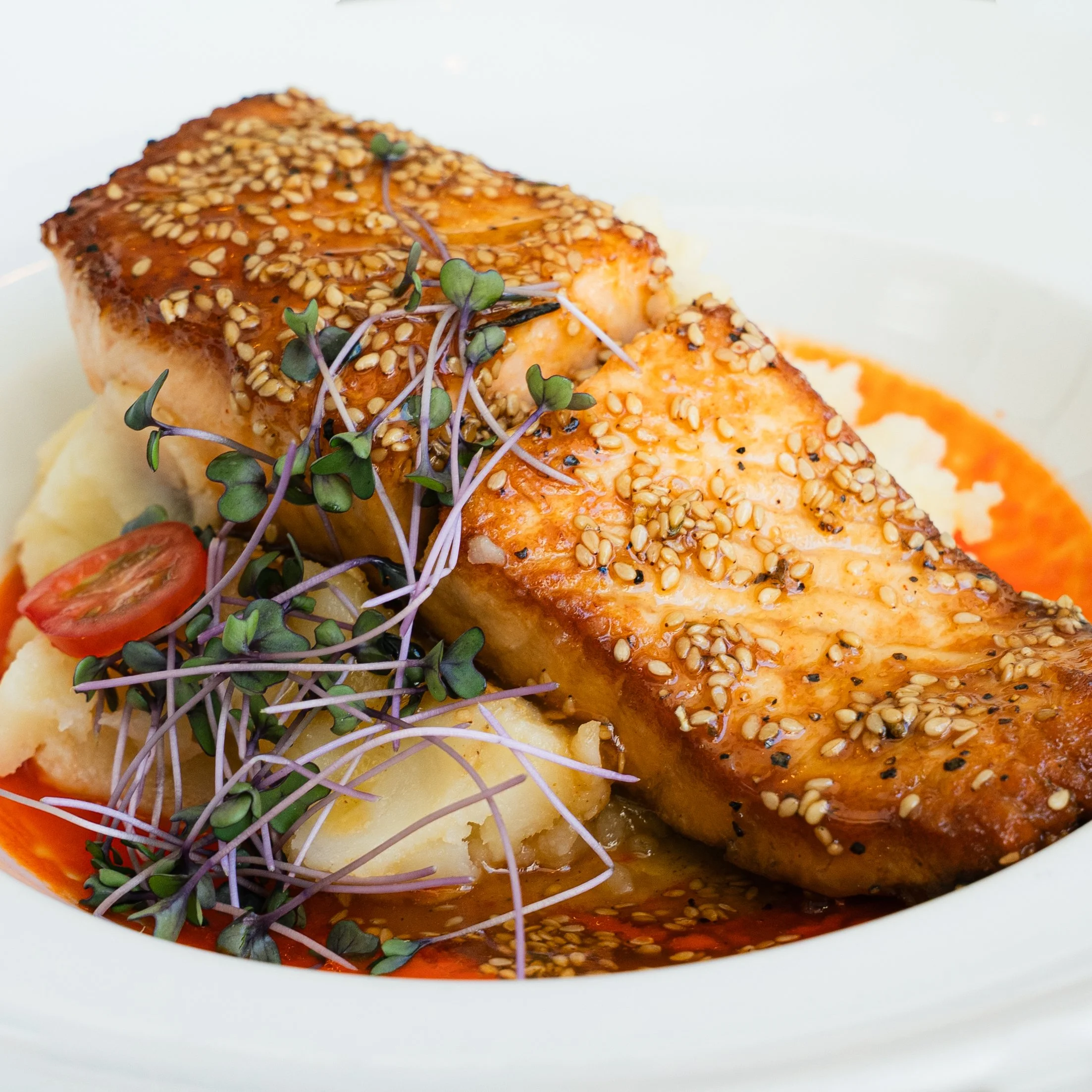 Close-up image of grilled salmon fillet topped with sesame seeds and garnished with microgreens on a plate with mashed potatoes.