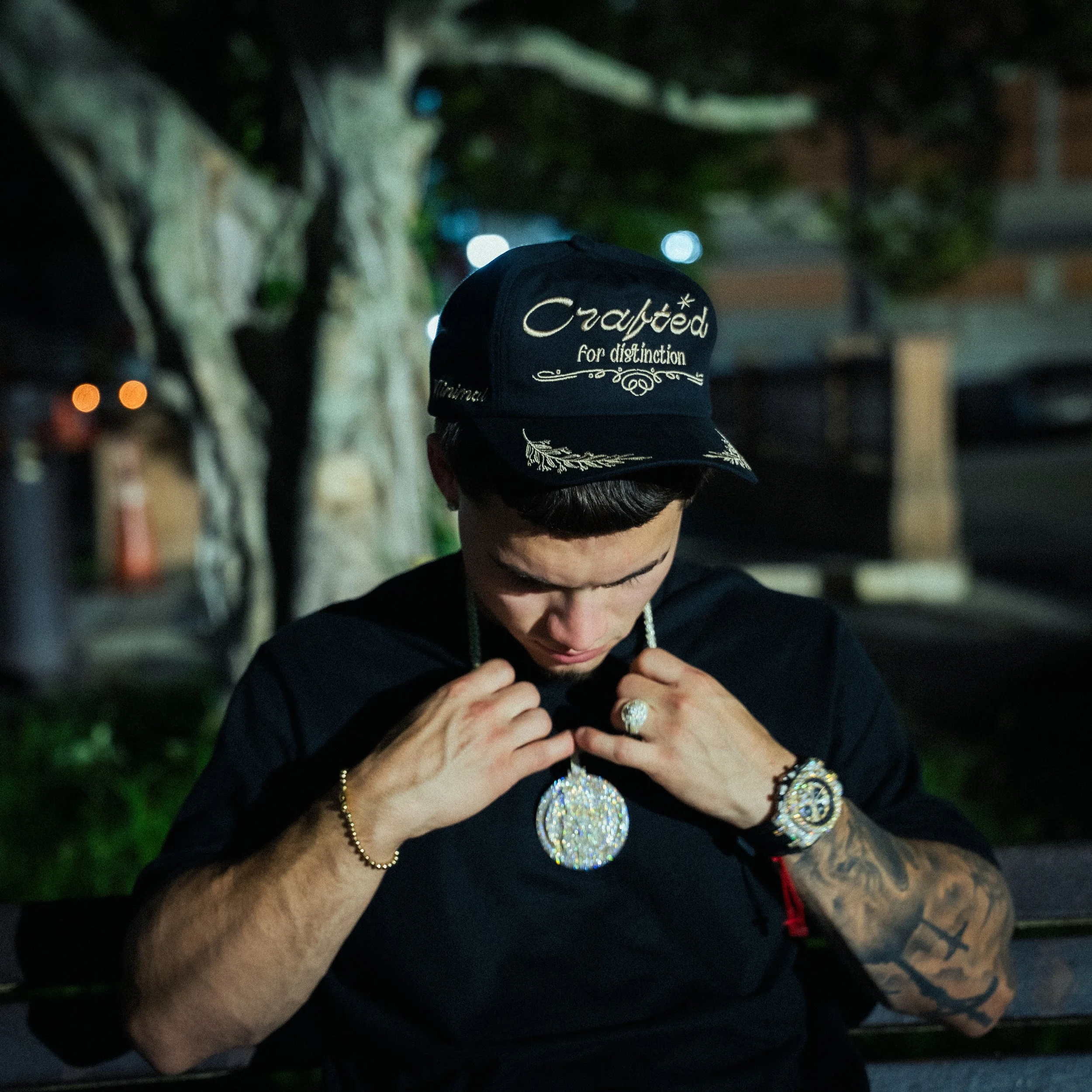 Persona usando una gorra negra con diseño y palabras brillantes, varias pulseras y anillos, en un entorno oscuro con iluminación de noche, algunas luces en el fondo.