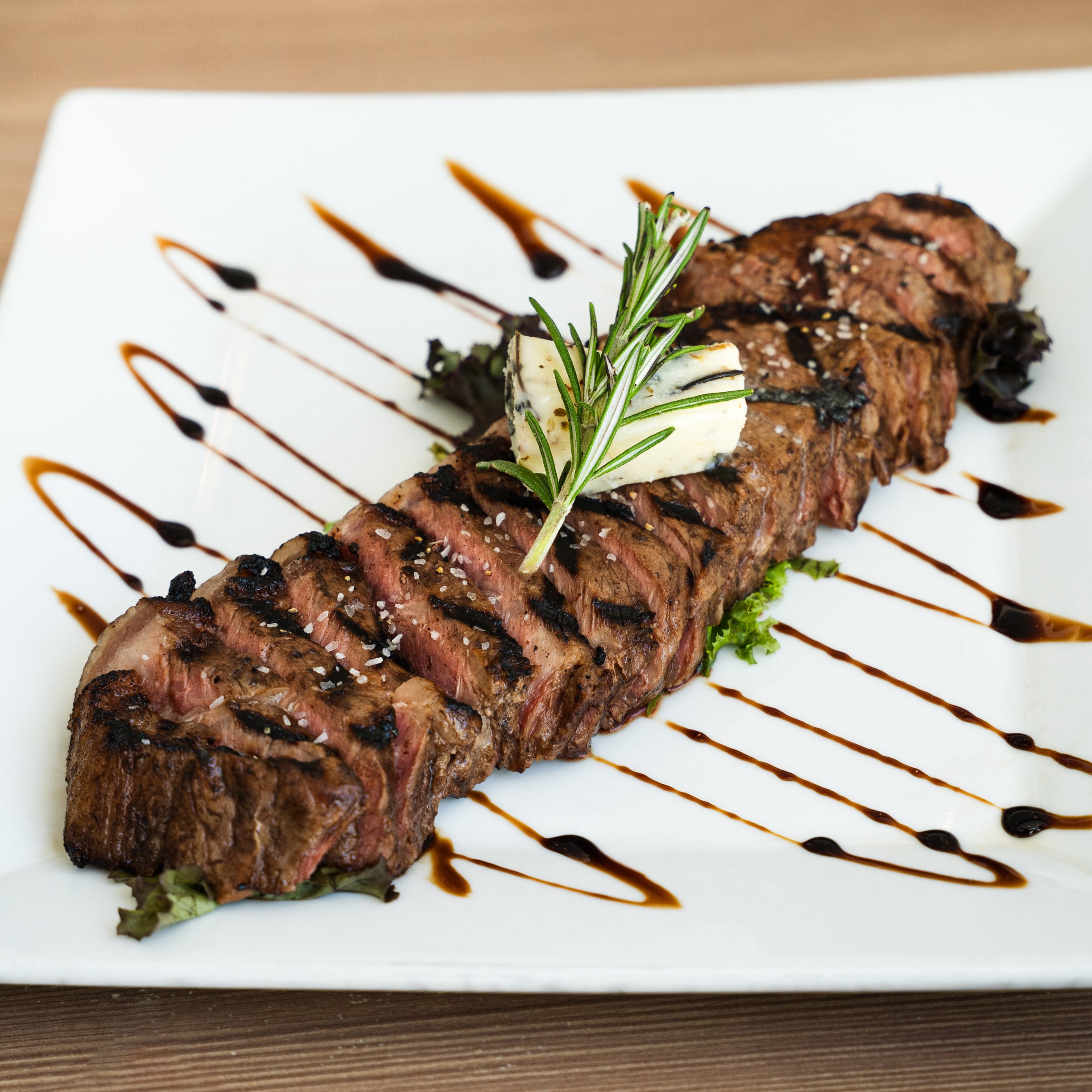 Grilled steak steak topped with rosemary, accompanied by a pat of herb butter, served on a white plate with decorative sauces.