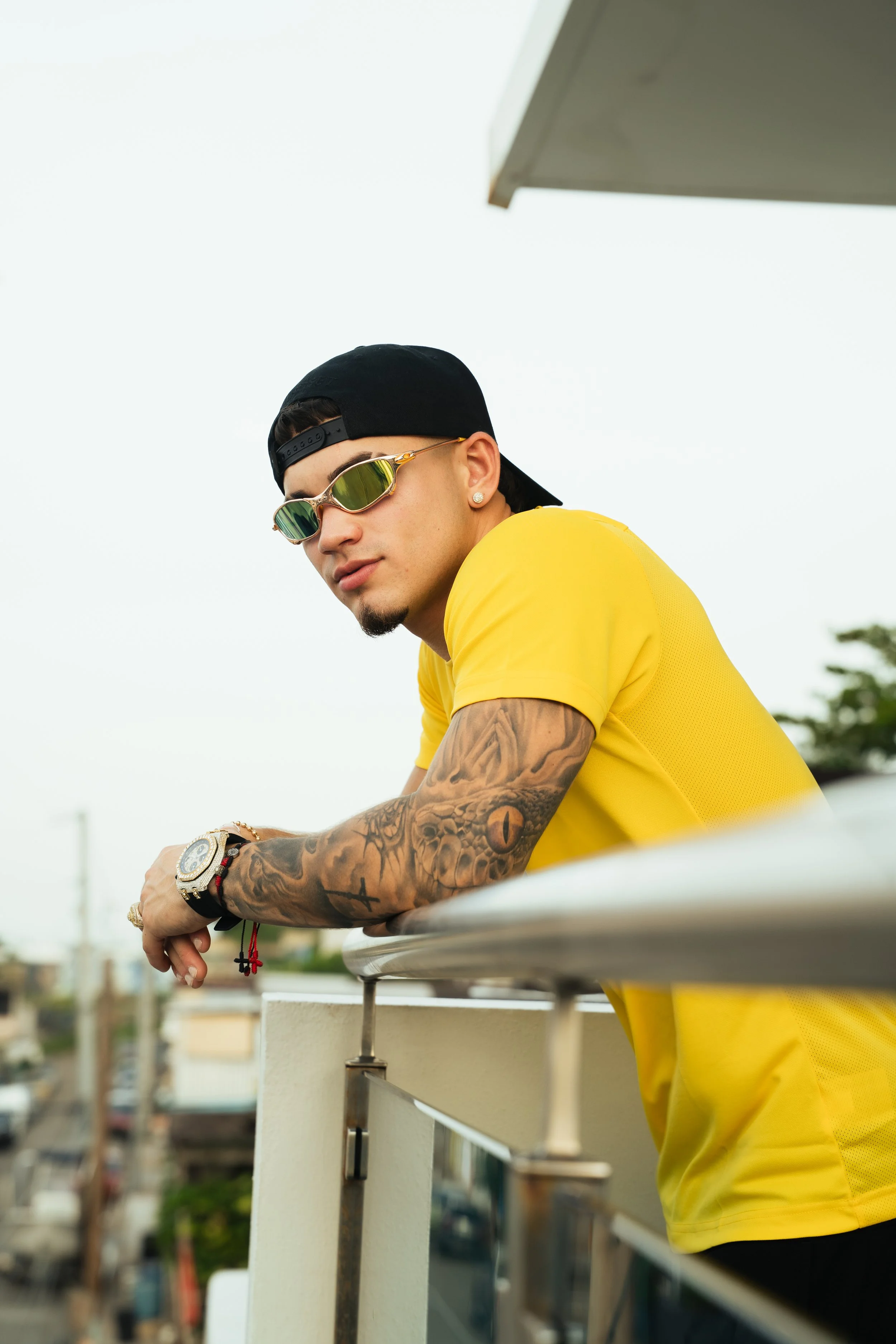 A young man wearing sunglasses, a black backward cap, a yellow t-shirt, and with tattooed arms leaning on a white railing on a balcony, with a blurred cityscape background.