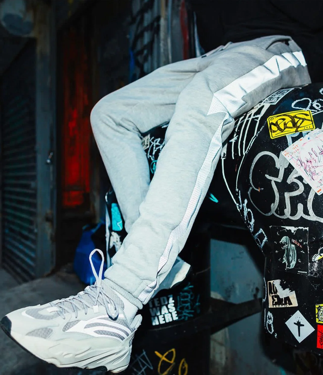 A person wearing light gray sweatpants and white athletic shoes sitting on a overturned skateboard decorated with stickers.